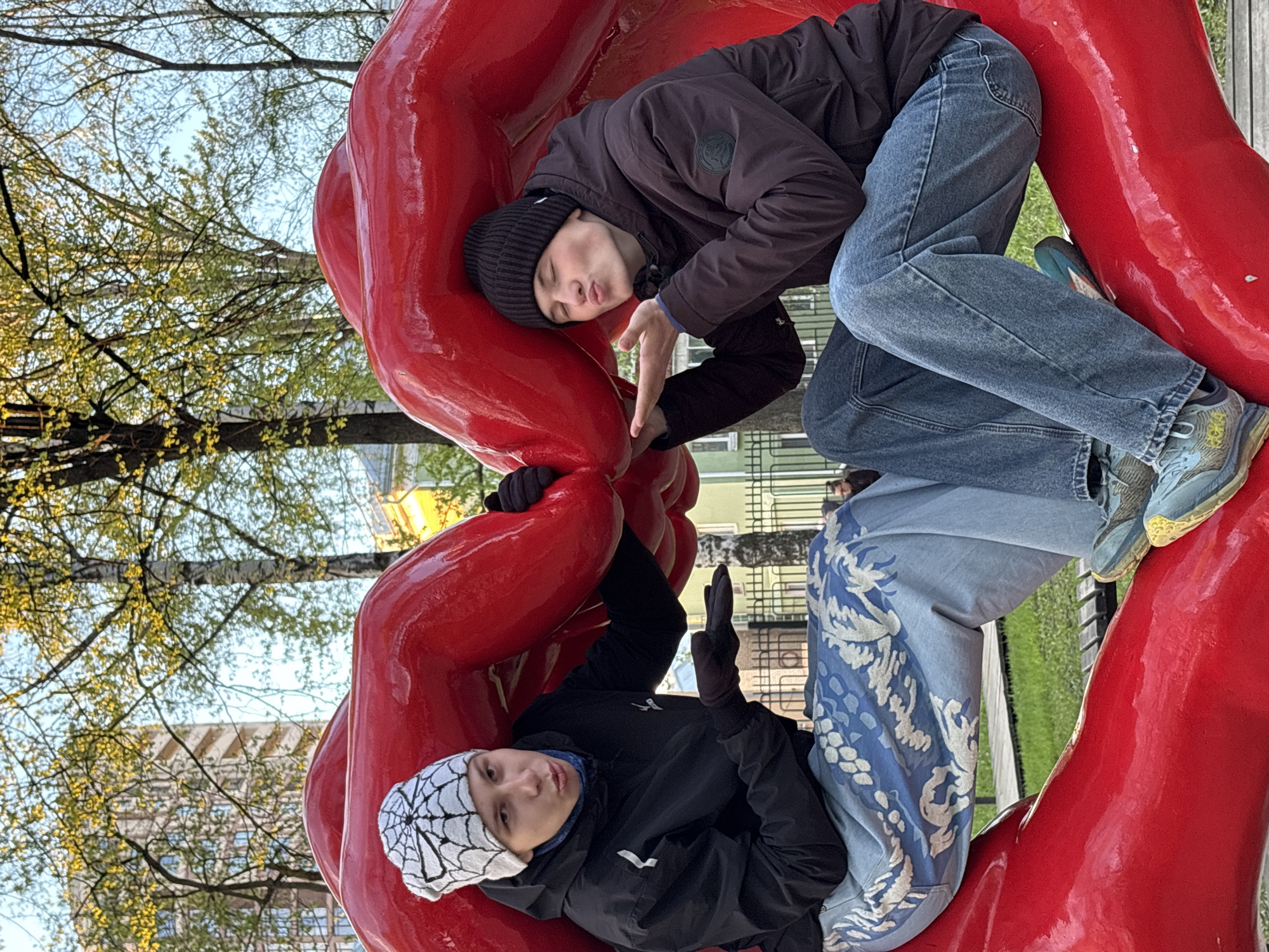 Two people are lying within the public art installation "Permyak – Salty Ears" in Perm, Russia. The sculpture consists of a large red circle with cutouts for two heads. One individual wears a black beanie, black jacket, and blue jeans. The other wears a white beanie, black jacket, and blue patterned pants. Trees and buildings can be seen in the background, suggesting an urban park setting.