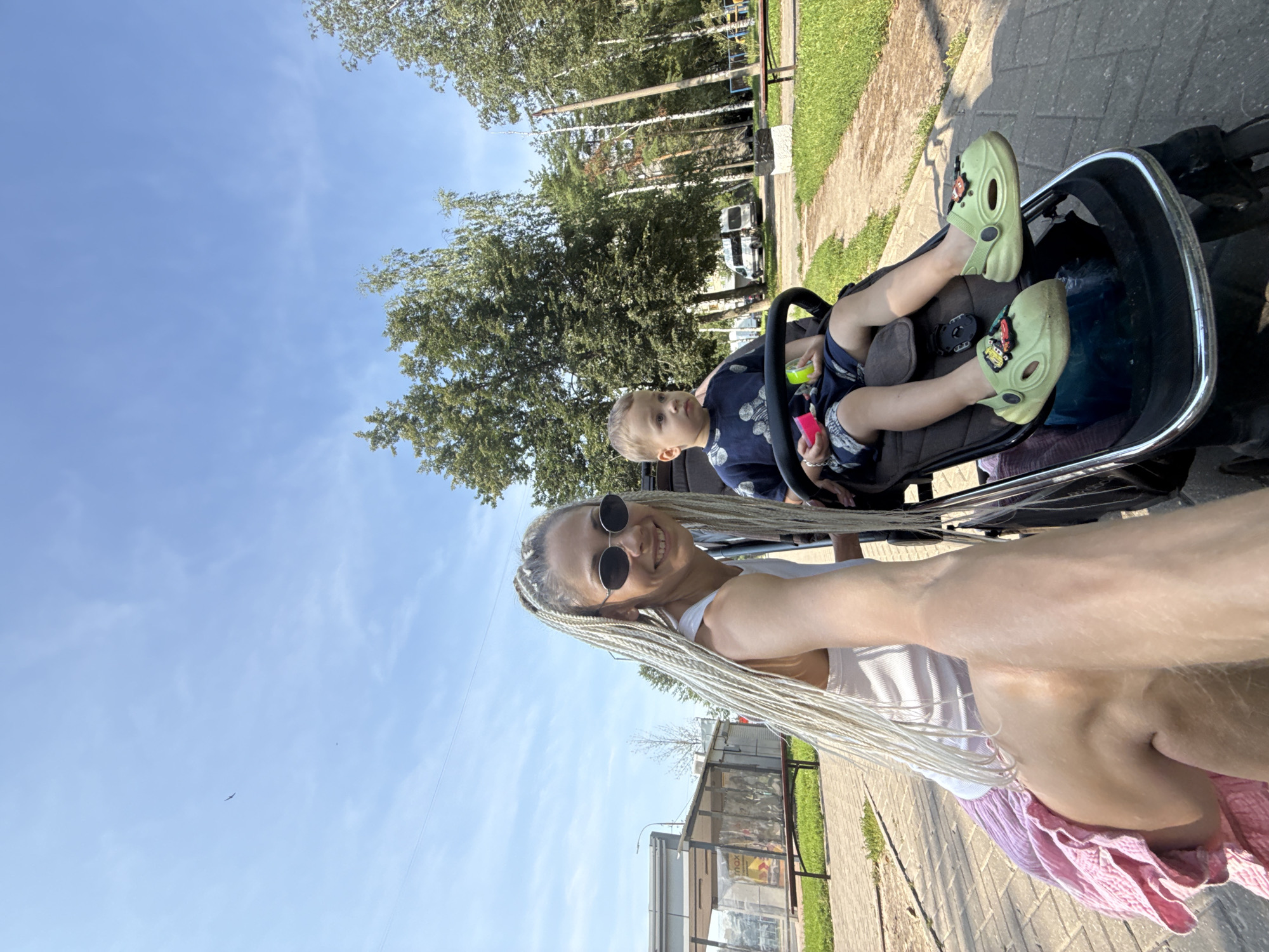 In the city of Rusanovo, Russia, a woman with long blonde braids and sunglasses smiles at the camera, holding the handle of a stroller. A young boy sits in the stroller, wearing a dark blue shirt and shorts, with green shoes adorned with car charms. The scene takes place outdoors on a paved path, with a grassy area and trees in the background. A bus stop shelter is visible to the left. The sky is clear blue with some wispy clouds.