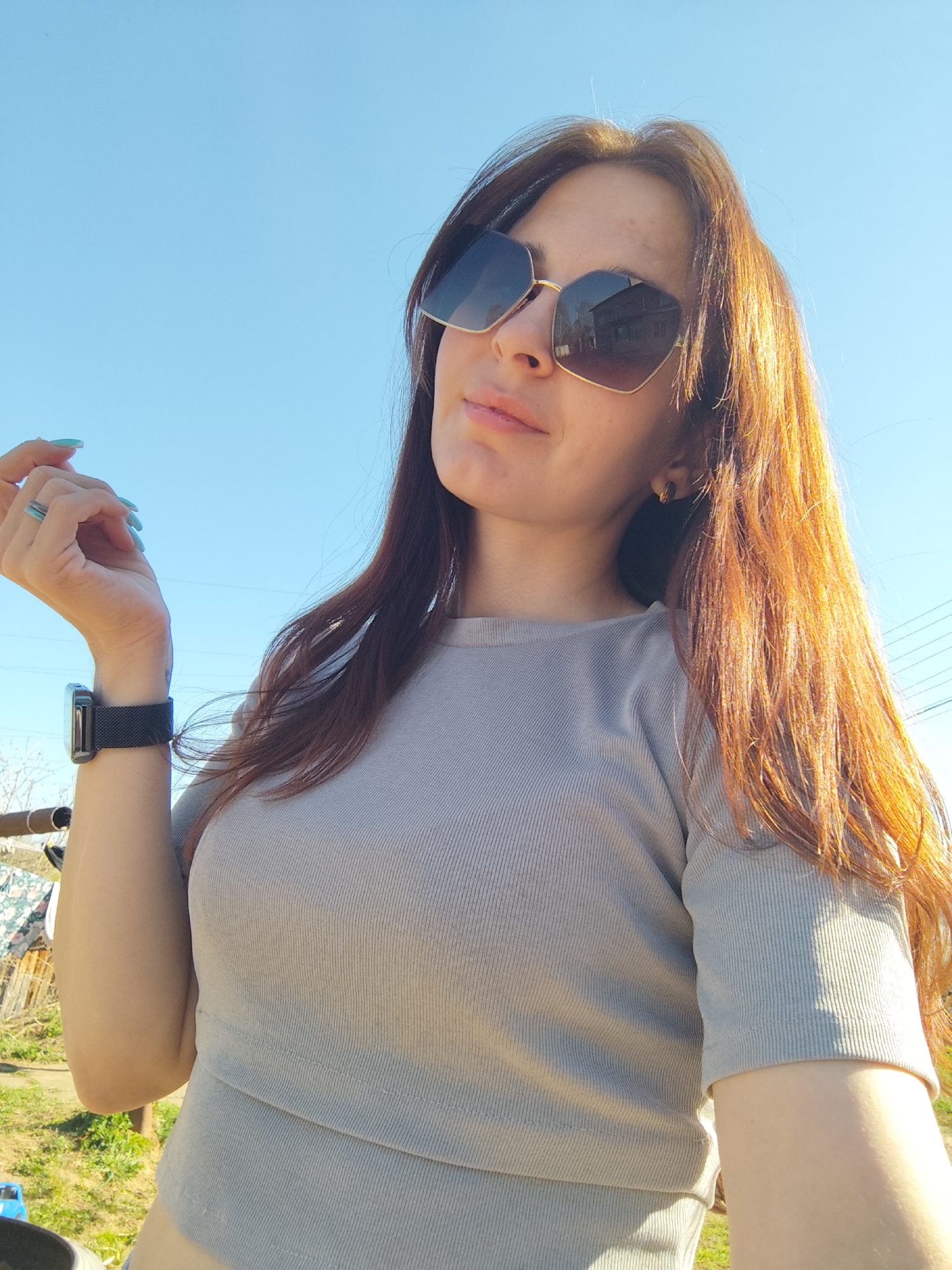 A woman with auburn hair is posing for a photograph outdoors. She is wearing sunglasses and a gray, short-sleeved shirt. On her left wrist is a black smartwatch. She is holding her left hand up, revealing a blue ring and matching nail polish. The background features a bright blue sky and indistinct elements of a yard, including grass, a wooden structure, and some clothes drying. There is no evidence to confirm the location.