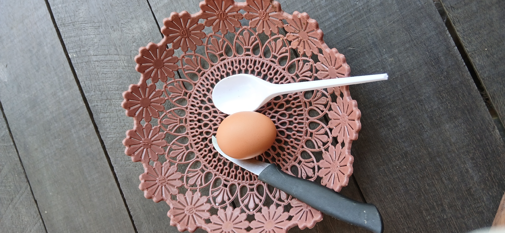An overhead shot shows a brown, circular plastic basket with a decorative, floral-like pattern, resting on a dark wooden surface. Inside the basket, an egg sits next to a white plastic spoon and a knife with a black handle. The basket is filled with a delicate, lace-like design. The wooden surface appears to be made of planks, possibly a table or deck, with visible grain and texture. The overall scene is dimly lit, suggesting an outdoor setting during daylight. This image was captured in Samarinda, Indonesia.