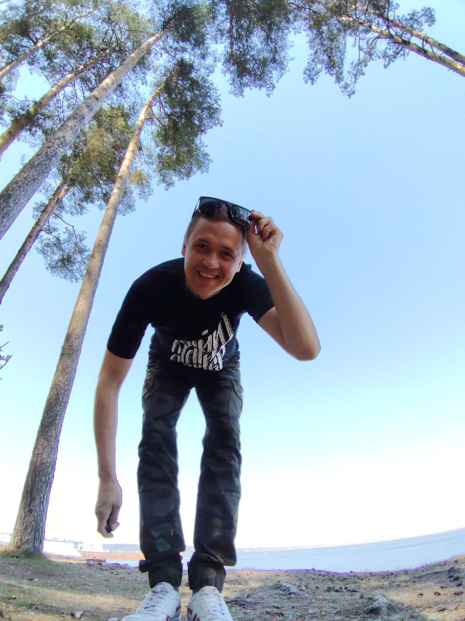 The image captures a smiling man from a low-angle perspective. He's wearing a black t-shirt with a graphic print, camouflage pants, and white sneakers. He holds sunglasses above his head with one hand. Behind him, tall trees stretch into a clear blue sky. The ground is covered in dried vegetation and pine needles, sloping slightly towards a body of water with land visible in the distance. The image was taken in the city of Chaikovsky, Russia.