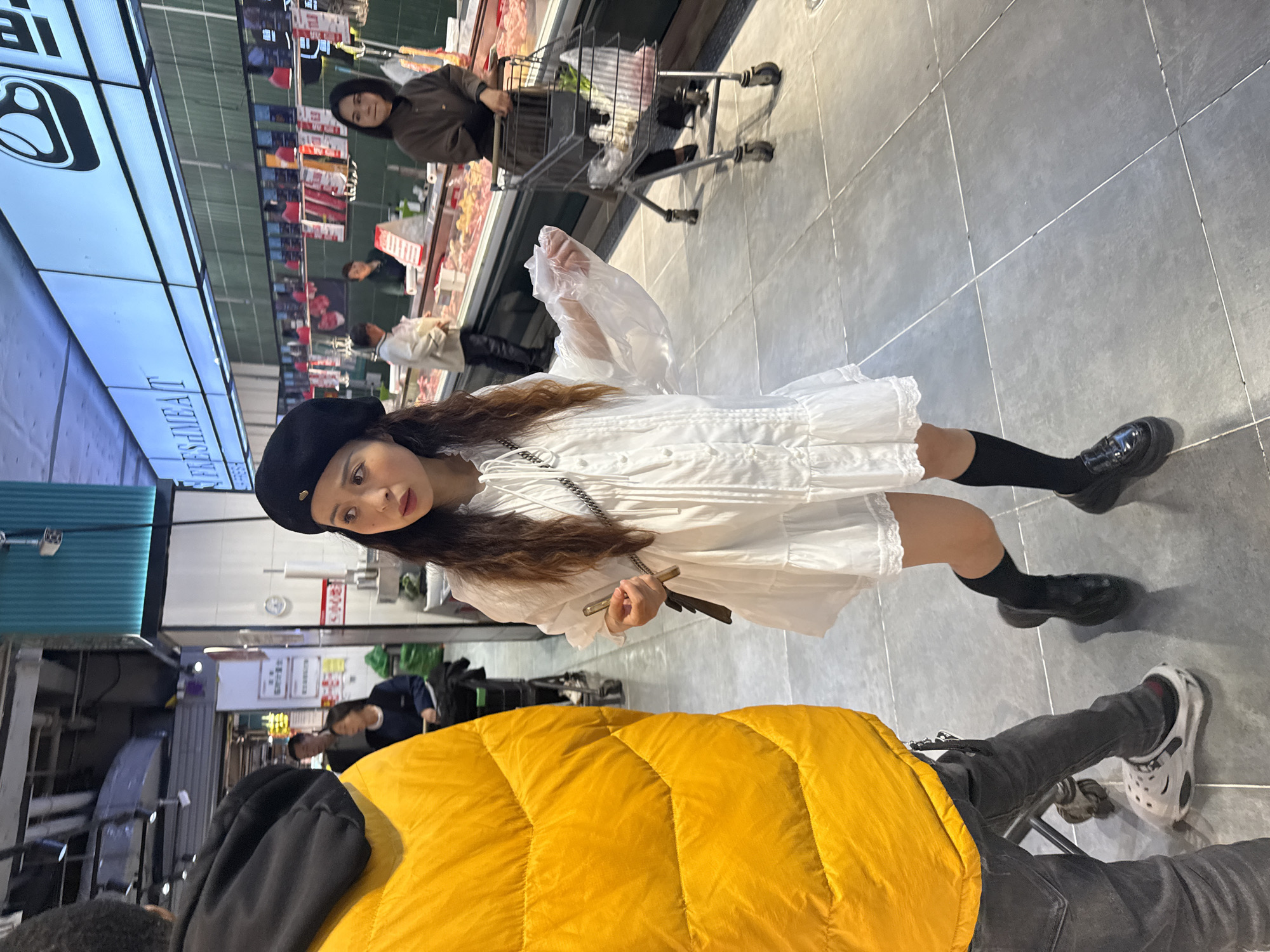 The image depicts a scene inside a supermarket. A young woman wearing a white dress, black beret, and black socks with shoes is holding a plastic bag. In the background, another woman is pushing a shopping cart. In the foreground, a person wearing a bright yellow jacket is visible from the shoulders down, accompanied by someone in gray pants and white Crocs. The setting appears to be a store with refrigerated meat products displayed behind the first woman. The location is likely Jiangjin, China, based on image metadata (not visible).