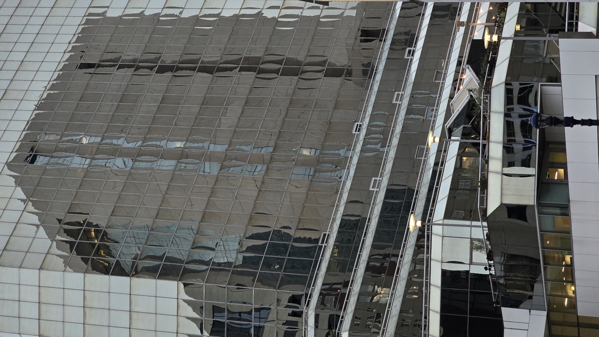 The image is a close-up, eye-level, full exterior shot of a modern glass-fronted building, reflecting another building. The building appears to be an office building. The facade is made of numerous glass panes, reflecting the distorted image of an adjacent building. The building has multiple levels with glass balconies and windows. The lighting is soft, suggesting it's either early morning or late evening. The sky is not visible in the frame. A streetlamp is visible in the foreground. The scene is devoid of people. The building is located in the city of San Diego, California, United States of America.