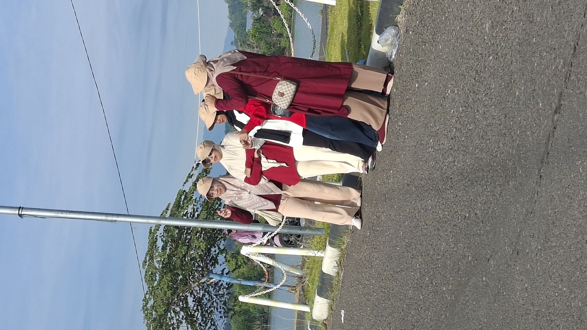 Four women, all wearing hats and loose-fitting clothing, stand in a line on the side of a road. The woman on the far left is wearing a beige hijab and a maroon top with tan pants. The second woman from the left is wearing a white hijab and a red top with white pants. The third woman from the left is wearing a black hijab and a red top with dark blue jeans. The woman on the far right is wearing a beige hijab and a maroon top with tan pants. They are all facing towards the right of the frame. A metal pole with a thin wire extending from it stands to the left of the women. Behind the women is a body of water, a white fence, and trees. The scene appears to be outdoors on a sunny day. The location is identified as Lhokseumawe, Indonesia.