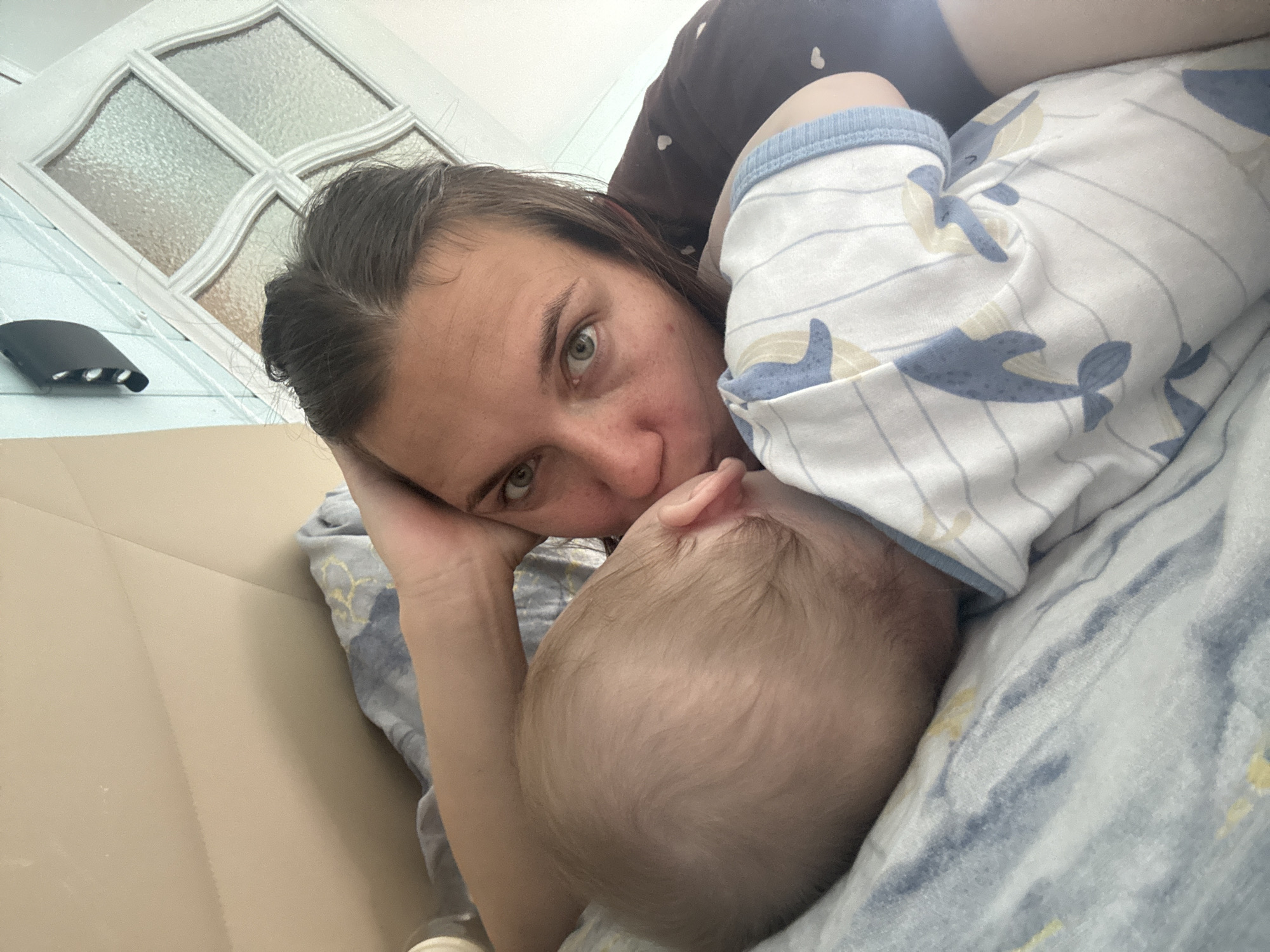 A woman with dark hair and green eyes is lying on a light blue and white patterned bedspread next to a baby. The baby is wearing a white onesie with a blue whale pattern. The woman's face is close to the baby's head, and she appears to be looking directly at the camera. Her hand is resting on the bedspread near the baby. A white door with a textured glass panel is visible in the background. A black object is attached to the door. The scene is indoors, likely in a residential setting. There is no verifiable evidence that this photo was taken in Razdolie, Russia.