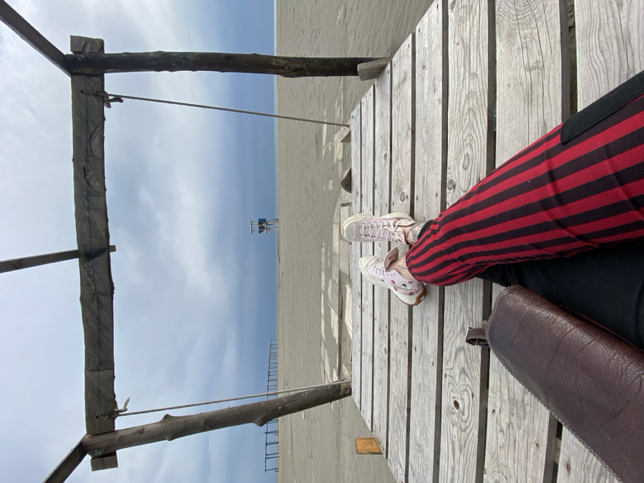 The image depicts a first-person view from a weathered wooden platform on a sandy beach in Ulcinj, Montenegro, under an overcast sky. In the immediate foreground, a person's legs are extended, clad in red pants with vertical black stripes and light pink high-top sneakers. A dark brown, textured bag rests on the left thigh. A rudimentary wooden frame structure, consisting of vertical posts and a horizontal crossbeam, partially frames the upper left of the scene, with a rope hanging from the left post. Beyond the platform, a wide expanse of light brown sand stretches towards the horizon. A blue and white lifeguard tower stands centrally on the distant beach, slightly to the right. Further to the left on the sand, a linear series of dark wooden posts connected by crossbeams is visible. The background consists of a calm, dark blue-grey sea meeting a light grey, heavily clouded sky.