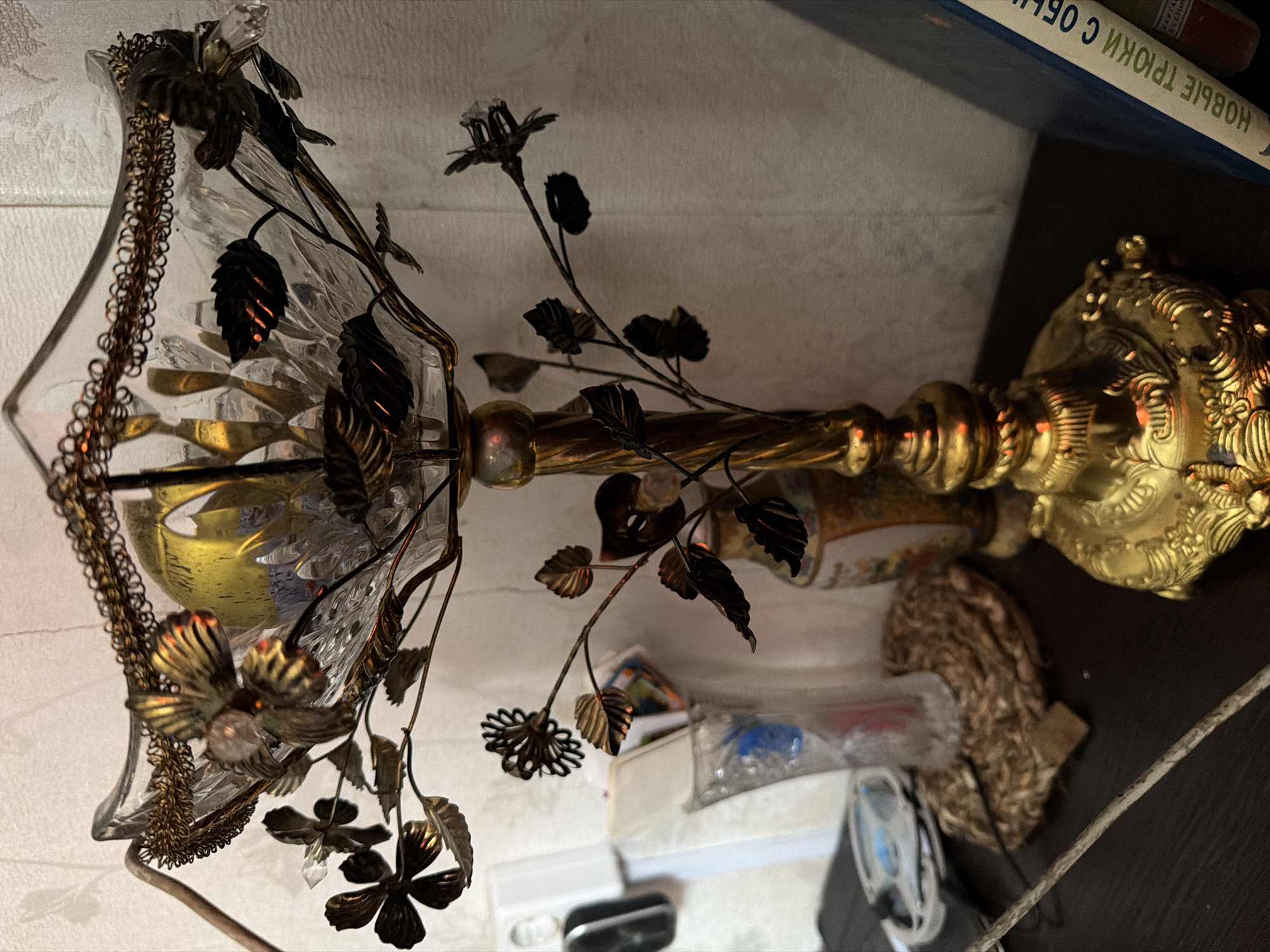 The image shows an ornate, antique-style lamp with a glass shade and decorative metal flowers and leaves. The lamp is golden and appears to be made of metal and glass. It is positioned on a dark surface, possibly a table or shelf, with a textured, light-colored background behind it. Several small objects are scattered around the base of the lamp, including what appears to be packaging, some plastic items, and a piece of woven material. In the upper right corner of the image, a book with Russian text is visible. The text on the book appears to read "НОВЫЕ ТРИОКИ С ОБ" which translates to "NEW TRIOKI WITH OB". The lighting in the image is warm and somewhat dim, suggesting an indoor setting, possibly in the evening. The overall impression is of a vintage or decorative item placed in a domestic environment. There is no activity or event taking place, it is a still image of an object.