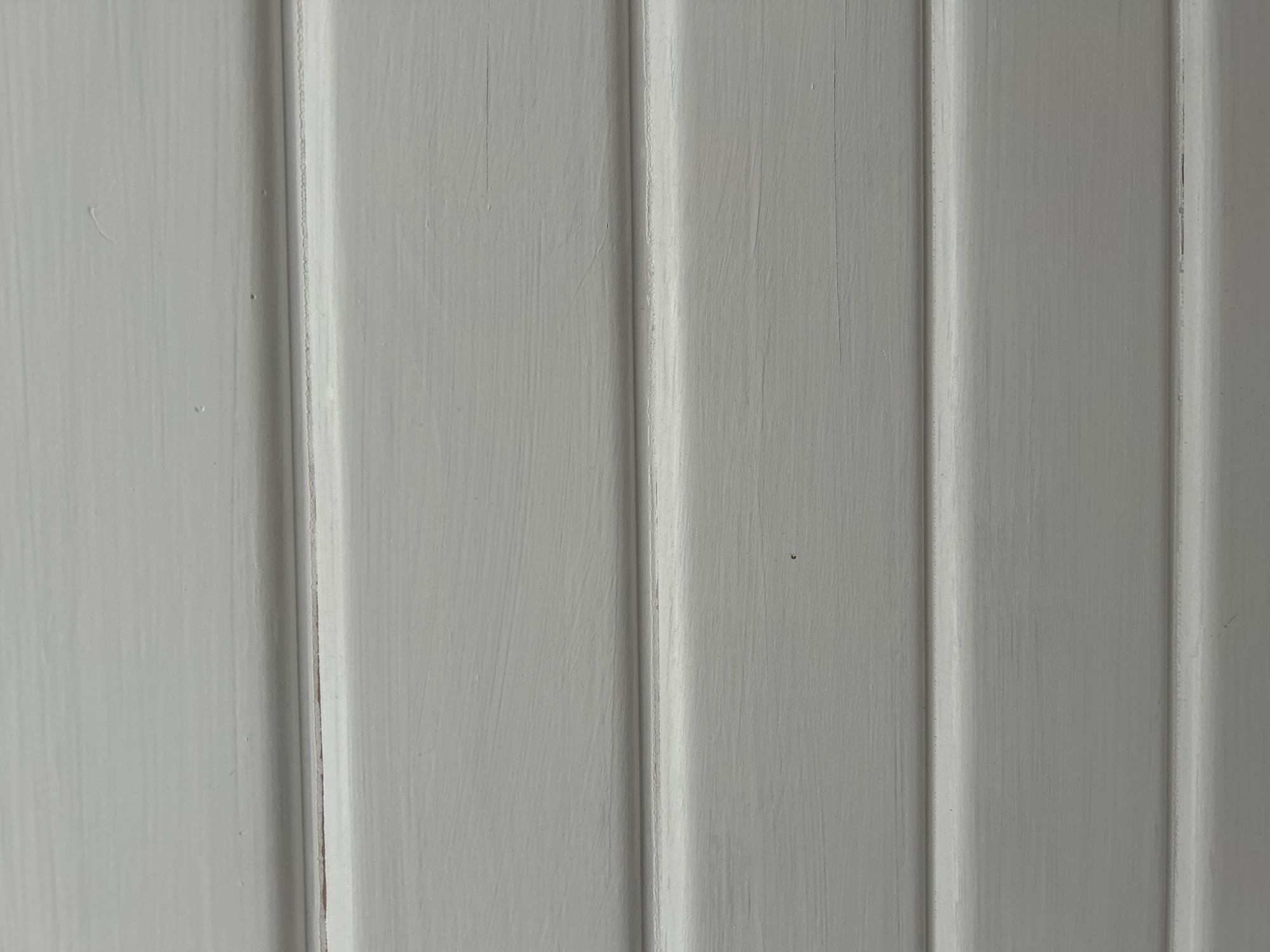 A close-up, overhead, full-frame shot shows a weathered, light gray painted wooden surface. The surface is composed of horizontal planks or boards, each with a slightly beveled edge that creates a subtle shadow line between them. The paint is applied unevenly, with visible brush strokes and some areas where the underlying wood grain peeks through, especially in the crevices between the boards. There are a few small imperfections on the surface, such as tiny nicks, scuffs, and what appear to be small holes or divots. The lighting is even and diffuse, with no strong shadows. The overall impression is one of a rustic or aged wooden material.