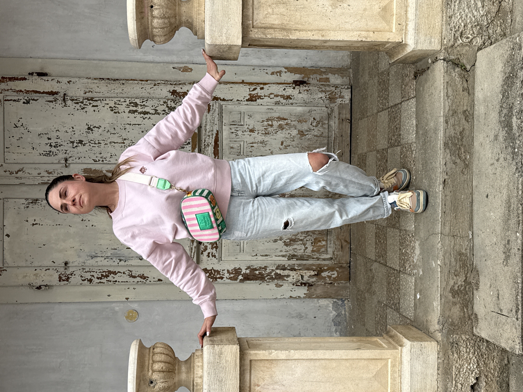 A woman is standing in a doorway. The location is Herceg Novi, Montenegro. The woman is wearing a pink sweatshirt, ripped light blue jeans, and tennis shoes, and she has a small striped bag slung over her shoulder. She has her right arm raised. She is standing on stone tile steps. The doorway has stone columns on either side and a weathered, white, peeling door in the background.