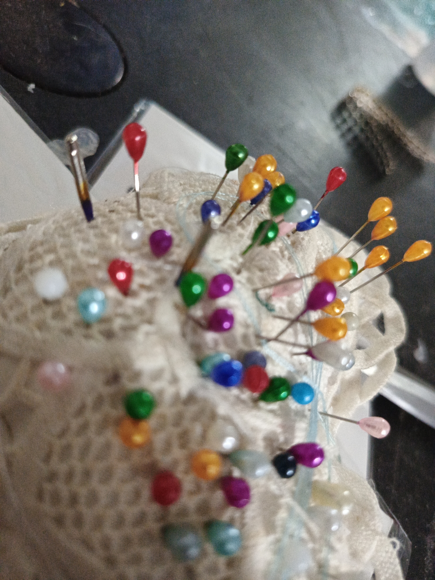 The image shows a close-up, slightly blurred view of a pincushion covered in various colored pins. The pincushion appears to be made of a light-colored, textured fabric, possibly lace or crochet. The pins have small, teardrop-shaped heads in a variety of colors, including red, green, blue, yellow, orange, purple, pink, and white. Some of the pins are angled, while others are more upright. The background is out of focus but appears to be a dark, textured surface. There are no people or identifiable landmarks visible in the image. The image does not contain any information that would allow for its location to be identified within the city of Juan Griego, Venezuela.