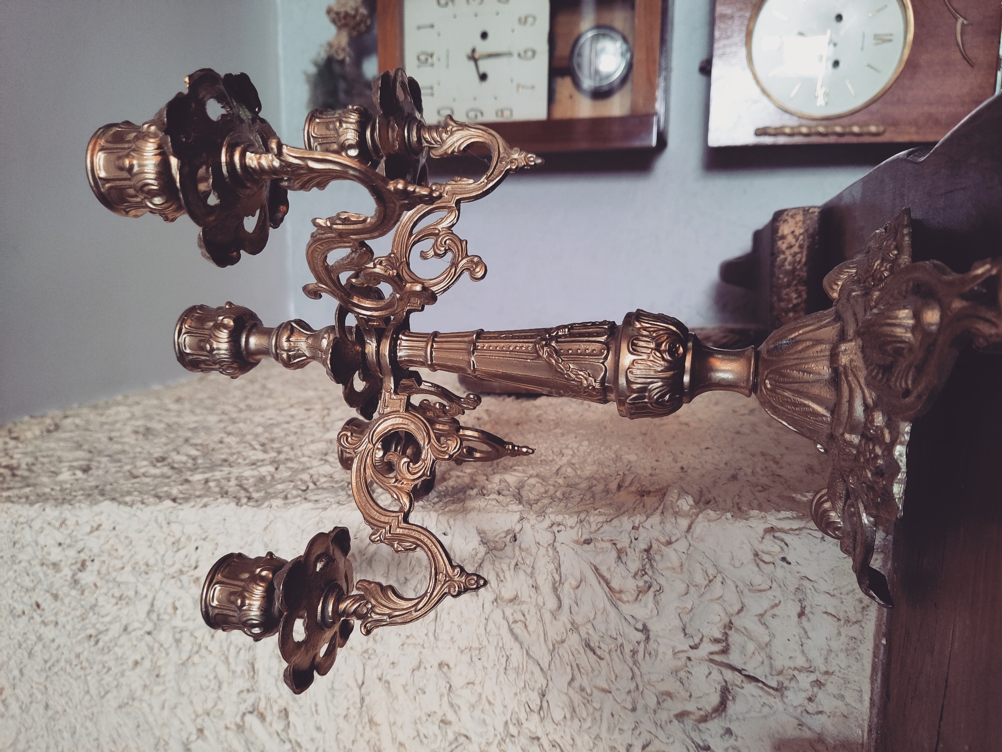 This is a close-up, eye-level, medium shot of a decorative, antique brass candelabrum. The candelabrum is ornate, with multiple arms and candle holders, and rests on a wooden surface. The background features a textured, off-white wall and a partially visible wooden clock with Roman numerals. The lighting is soft, with the candelabrum casting a slight shadow. The image appears to be taken indoors, in a dimly lit room. The location is the city of Norviliškės, Lithuania. There are no people or animals visible in the image.