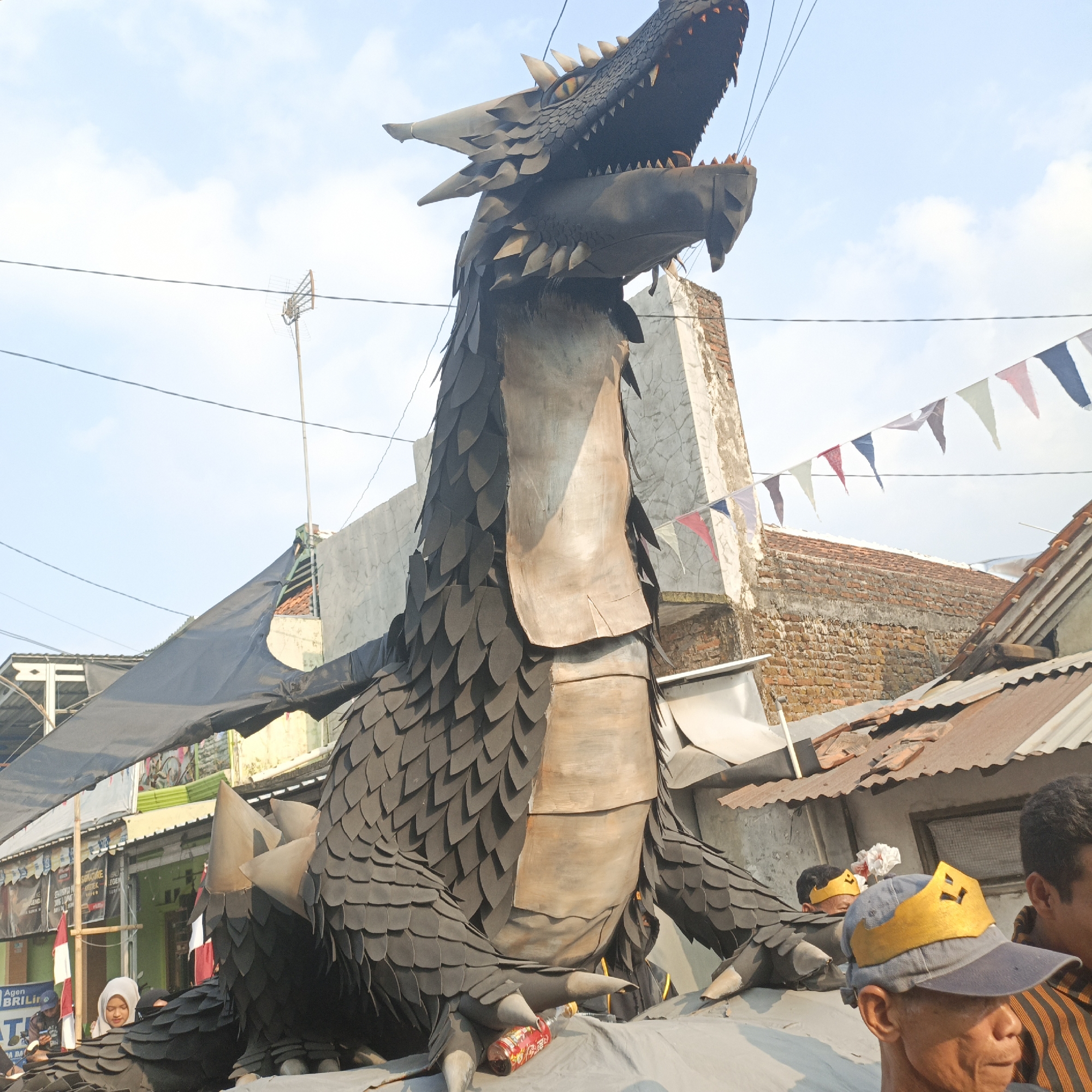 This is a photograph taken outdoors in the city of Tanahbaya, Indonesia, likely during a daytime celebration or parade. The main subject is a large, dark-colored dragon effigy, appearing to be constructed from layered materials resembling scales. Its head is raised with its mouth open, revealing sharp teeth and yellow eyes. The effigy is positioned on a raised platform, partially covered with gray fabric.
In the foreground, several people are visible. One man in a blue baseball cap with a yellow crown-like decoration is looking down. To his right, another man with dark curly hair and a striped shirt is partially visible. Behind the dragon effigy, a woman wearing a white hijab is looking towards the left. Further in the background, there are buildings with brick and concrete walls, corrugated metal roofs, and electrical wires crisscrossing overhead. Colorful triangular flags are strung between buildings. A red and white Indonesian flag is also visible hanging from a pole. The sky above is bright with some clouds. A bottle with a red label is lying on the gray fabric in front of the dragon's claws.
