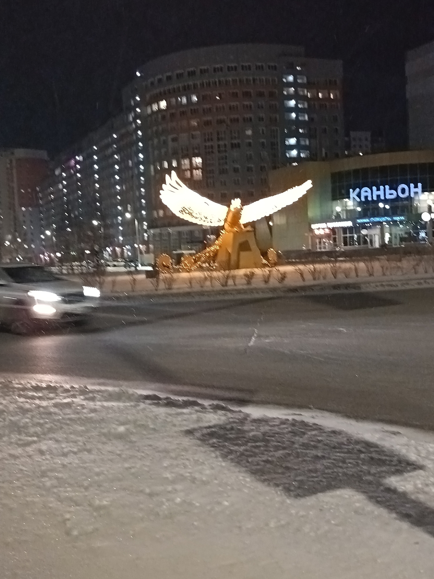 At night in Nizhnevartovsk, Russia, a lighted statue of an eagle with spread wings stands on a traffic island. In the background are multi-story buildings with lit windows and a building with the illuminated Cyrillic word "КАНЬОН". A car with headlights on is blurred, moving past the statue. The foreground shows a snow-covered road with dark patches.