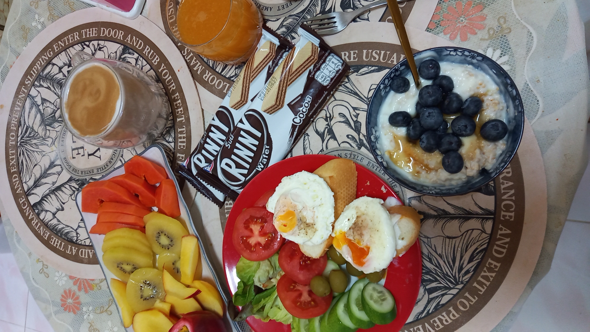 The image displays a breakfast spread on a table, featuring a variety of food items. In the foreground, a bowl of oatmeal topped with blueberries and drizzled with honey is visible. To the left, a red plate holds a salad with lettuce, tomatoes, cucumber slices, and olives, accompanied by two open-faced sandwiches topped with fried eggs. Beside the salad plate, there's a platter with sliced kiwi, papaya, and peach. In the background, a glass of coffee with latte art and a glass of orange juice are present. Two chocolate wafer bars are also on the table. The setting appears to be indoors, possibly a home or restaurant, with a decorative patterned tablecloth and placemats. The overall scene depicts a wholesome breakfast.