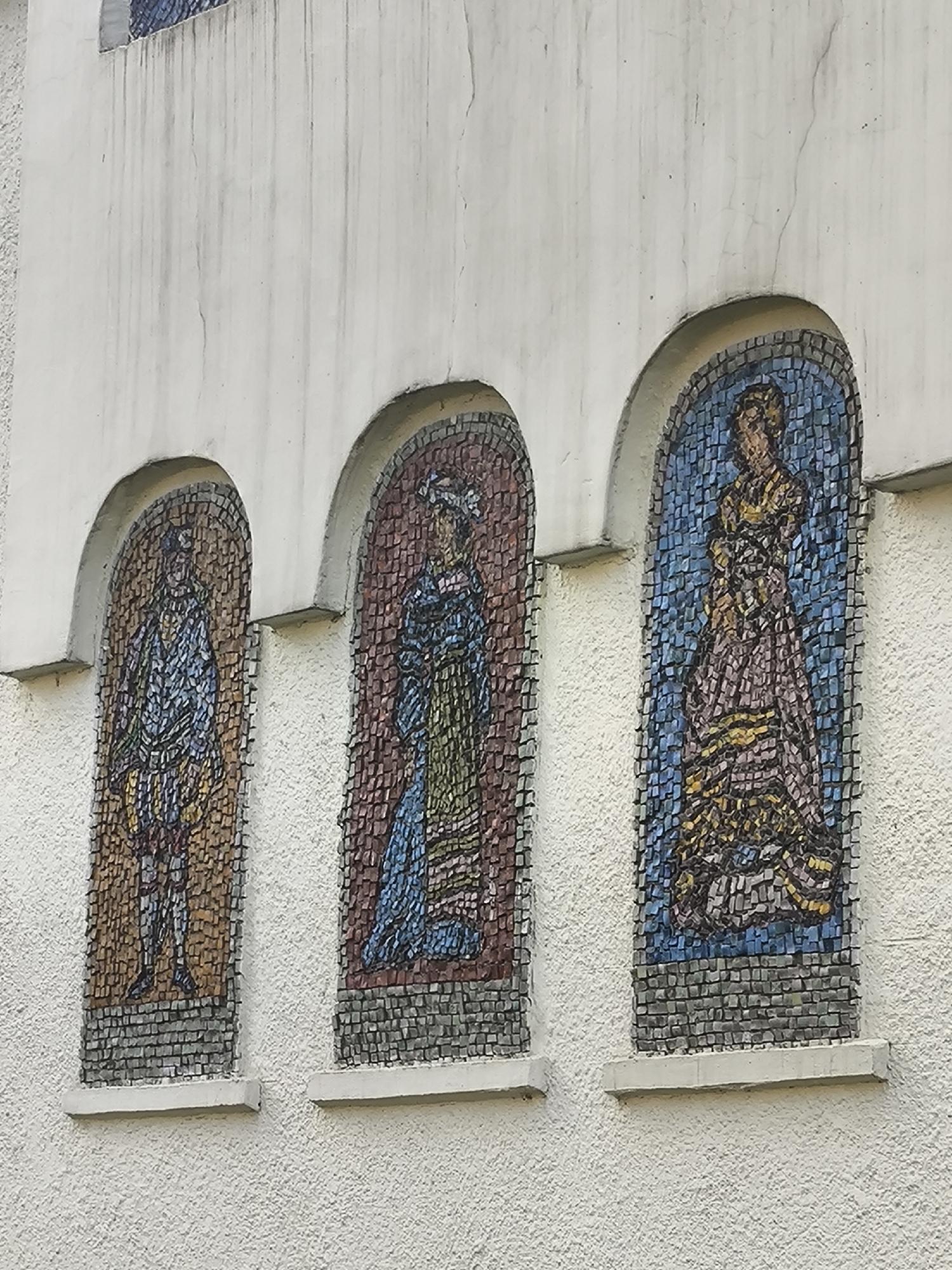 The image shows a section of a textured off-white building facade with three arched mosaic inlays. Each inlay features a stylized human figure. The leftmost mosaic depicts a figure wearing a patterned blue and yellow garment. The middle mosaic displays a figure in a blue dress with green and yellow accents. The rightmost mosaic shows a figure in a long, flowing brown and yellow dress against a blue background. The mosaics are set against the textured wall, with visible water stains running down from above the top edge of the arches. Each mosaic is framed by a darker mosaic border and sits above a concrete ledge. The overall scene appears to be an outdoor architectural detail.