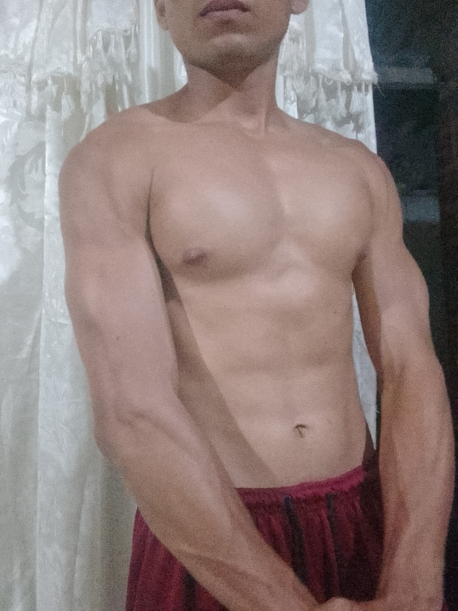 A torso of a man is centered in the frame. The man has brown skin, pectoral muscles, visible abdominal muscles, biceps, and forearms. He is wearing burgundy shorts. Behind him is a white curtain on the left, and a dark, undefined area on the right. There are no indications of the location being Juan Griego, Venezuela.
