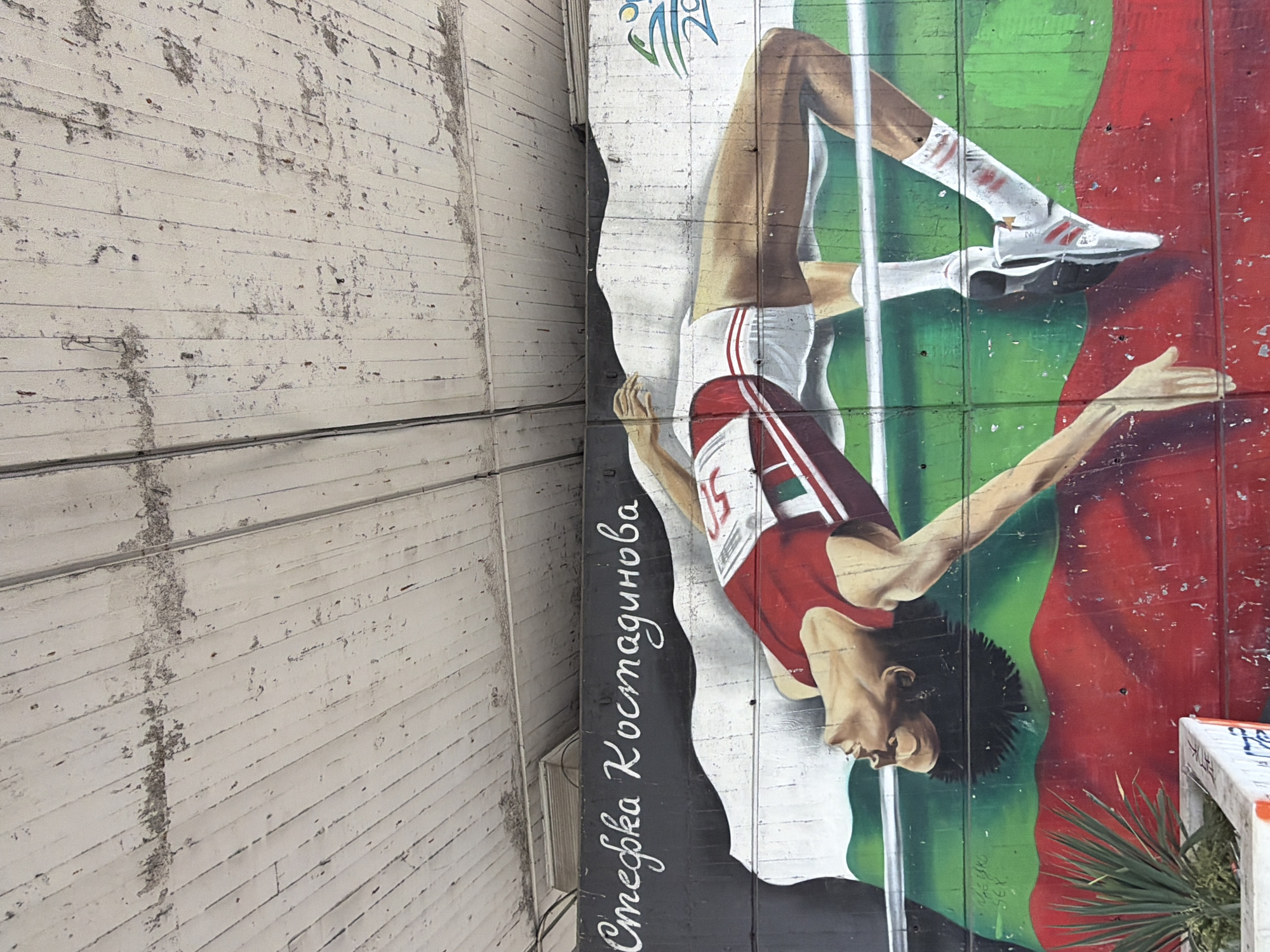 The image depicts a mural of Stefka Kostadinova on a building in Sofia, Bulgaria. The mural shows Kostadinova in action, high jumping with a red and white athletic uniform and the number 5 on her bib. Her hand and foot are extended towards the top of the painted high jump bar. The background behind her is colored in green and red. The text 'Stefka Kostadinova' is written vertically next to the painting. Adjacent to the painted wall is a building with a white wooden plank surface. A small palm tree is located at the bottom right of the mural.