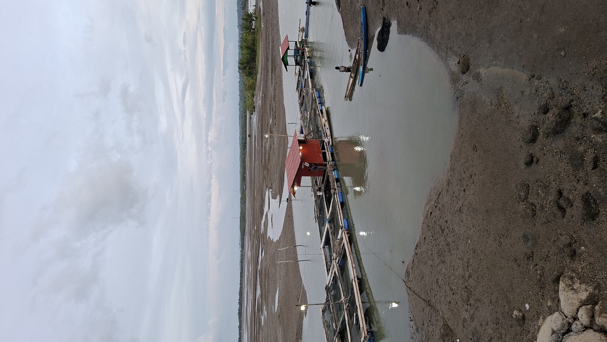 The image depicts a view of fish farms located in the waters near the shore of Lhokseumawe, Indonesia. The scene is characterized by a muddy, low-tide environment, with several floating structures serving as fish enclosures.
In the foreground, the muddy shoreline is visible, marked with rocks and debris. The water is calm and appears murky.
Dominating the midground are the fish farms, consisting of a series of interconnected floating platforms made of wood and blue barrels. On one of these platforms, a red, shed-like structure with a metal roof is present, with lights illuminating the area. A person can be seen standing near this structure. Further out on the water, another smaller, green-roofed structure sits on a similar floating platform.
Several boats are visible: a narrow wooden boat with a person standing in it is moored near the muddy bank, and a blue boat is partially submerged in the water. In the background, a person is seen near the green-roofed structure, and another person is in the water next to a boat.
The background shows a stretch of land with vegetation, possibly mangroves or palm trees, suggesting a coastal area. The sky is overcast with heavy clouds, indicating a diffused light condition, typical of dawn or dusk. The overall impression is one of an operational aquaculture site in a coastal environment.