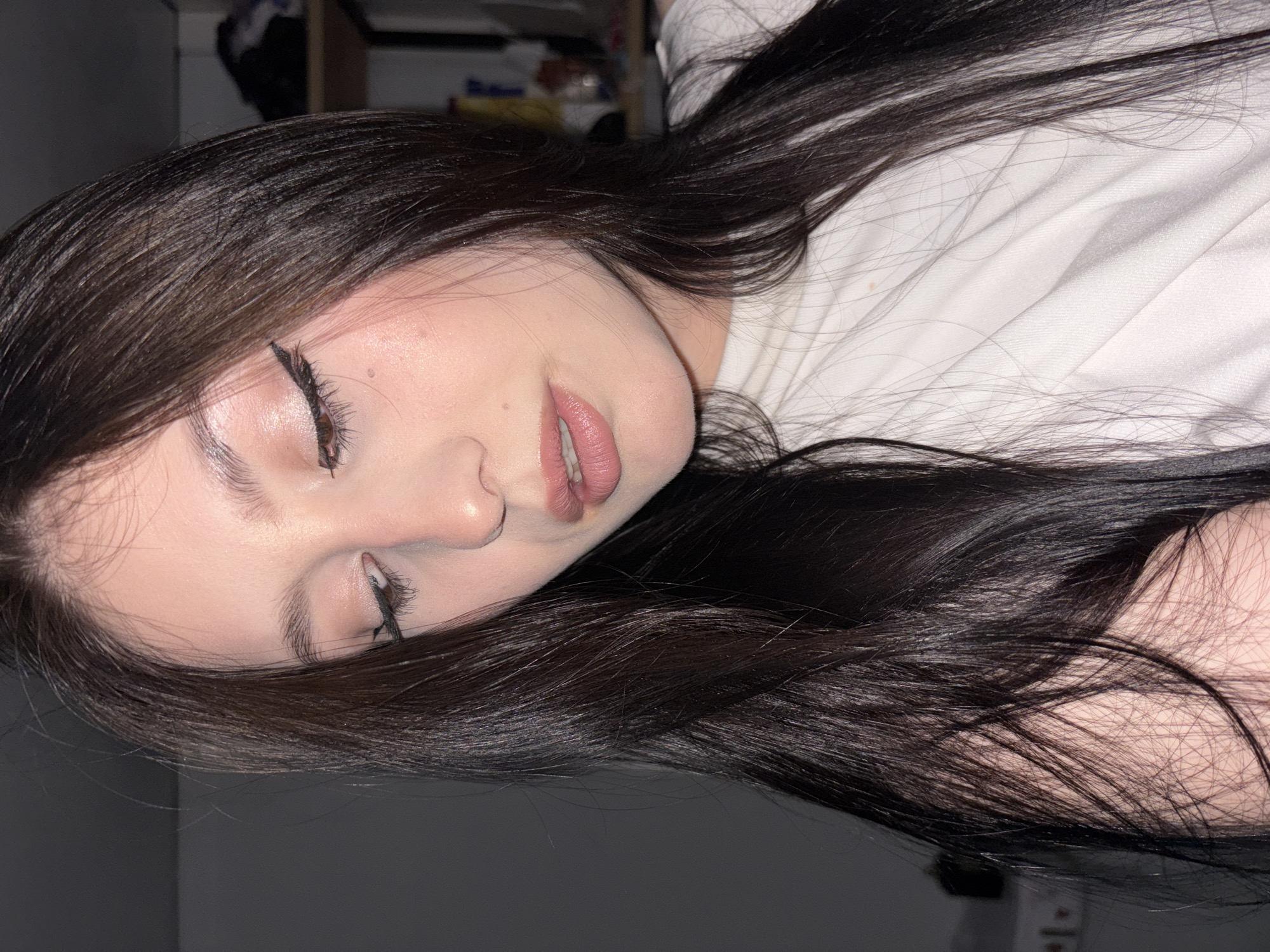 A young person, likely female, is depicted in a close-up portrait, lying down indoors. Their long, dark, straight hair is spread across a white surface, possibly a bed or pillow. They are wearing a white top. The subject's face is angled, with eyes partially closed and looking downwards, and lips slightly parted, conveying a relaxed or contemplative expression. Notable details include winged eyeliner, well-groomed eyebrows, and pinkish-nude lipstick. The skin appears smooth with subtle highlights. The lighting is soft and dim, suggesting an indoor setting during the evening or night. The background is mostly dark and out of focus, with some indistinct shapes visible in the upper left, indicating an enclosed space. No text is visible in the image. The provided location context is Фоки, Russia, though no specific visual cues confirm this.