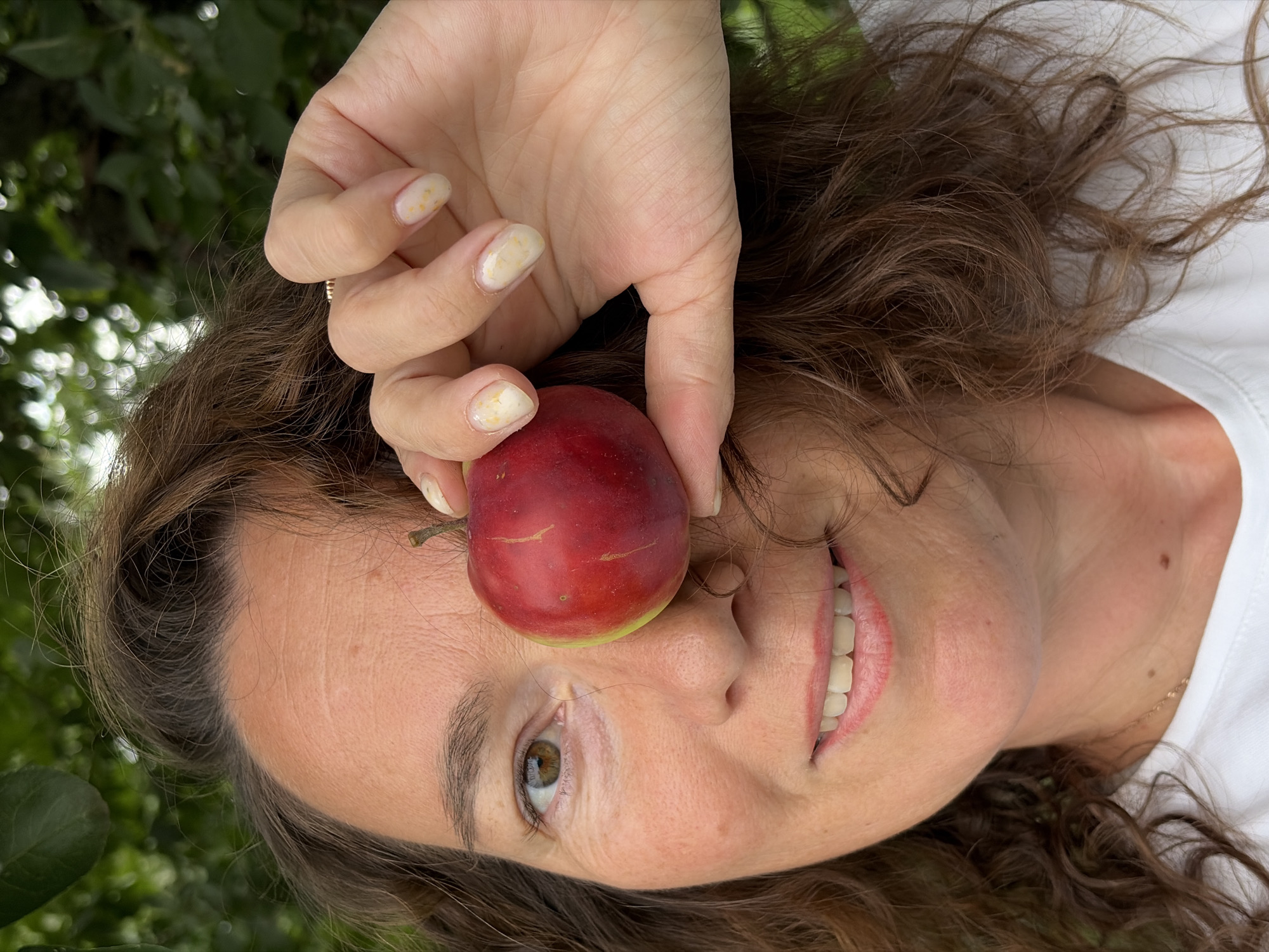 In Ольховка, Russia, a medium-shot portrait captures a woman with curly brown hair, her right eye obscured by a red apple she is holding in her right hand. Her left eye is visible, as is her smiling mouth and white t-shirt. The background is blurred green foliage.