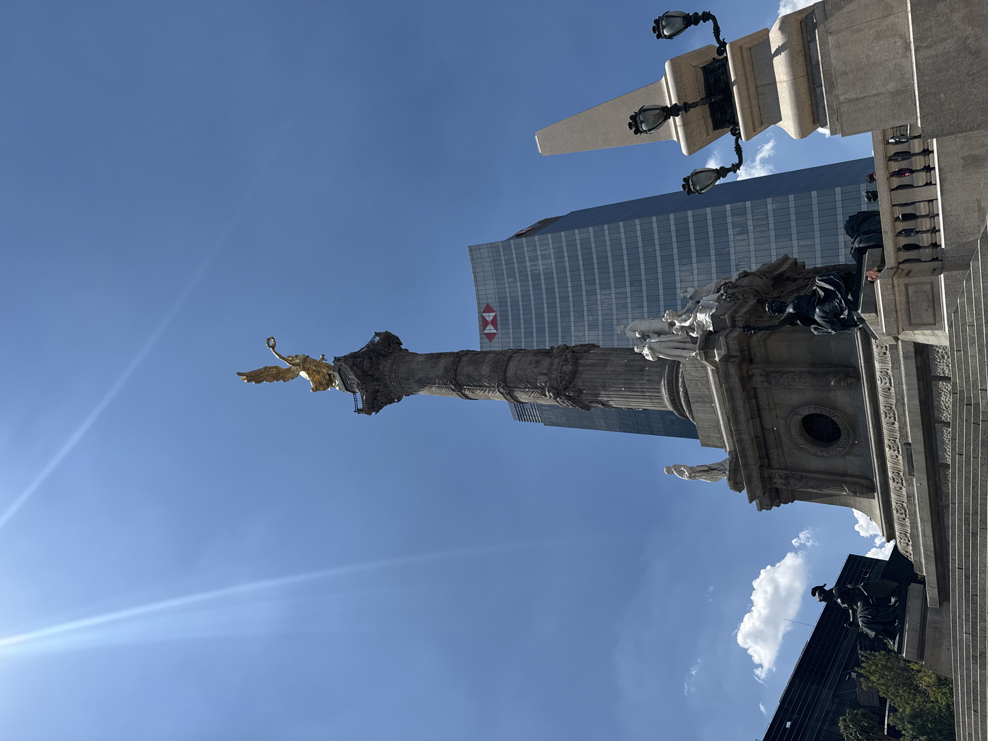 The image features a low-angle view of the Angel of Independence monument in Mexico City, Mexico. The golden angel statue is at the top of a tall column, with neoclassical sculptures at its base. In the background, a modern glass-and-steel skyscraper is visible, bearing the logo of HSBC bank. The sky is blue with a few scattered clouds. The image includes details of the surrounding architecture, such as lampposts and stairways, and the monument’s sculptures at the base.
