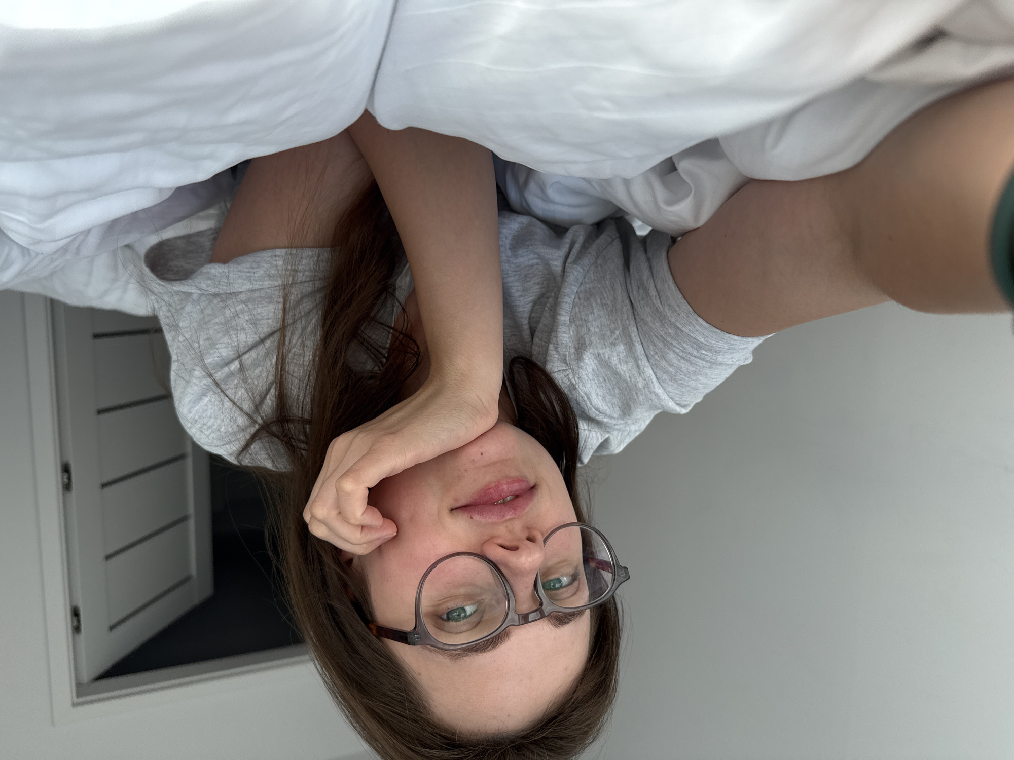 A young Caucasian woman with long, brown hair and glasses is lying on a white bed. She is wearing a grey t-shirt and her right hand is supporting her chin. The background shows a white wall and a partially open door with horizontal paneling. The lighting is soft and natural, suggesting it is daytime. The scene appears to be indoors, likely a bedroom. The location is not identifiable as Szklarska Poręba, Poland based on the visual information provided.