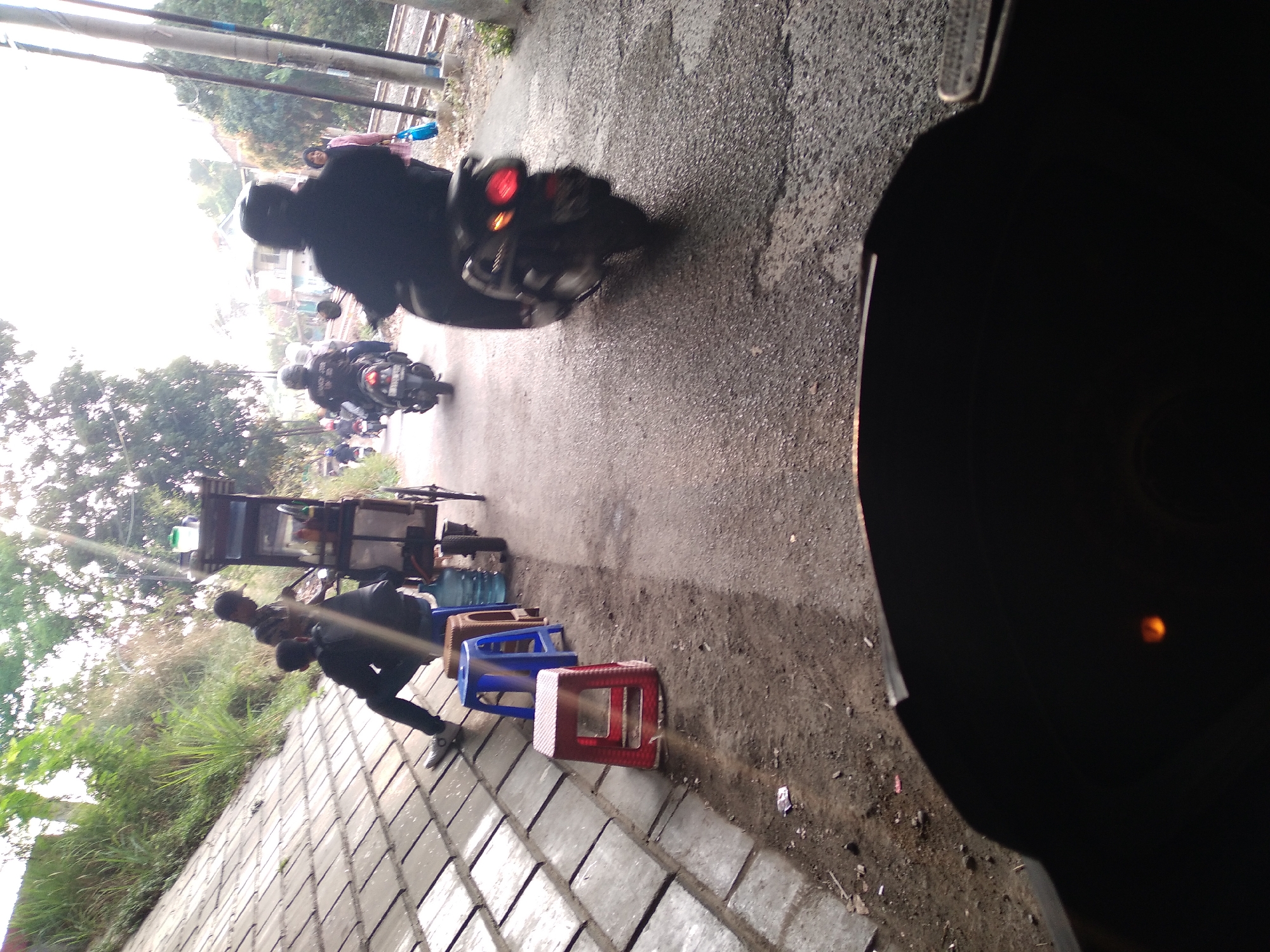 The image captures a street scene in Cimahi, Indonesia, likely near a railway line based on the presence of tracks in the background. In the foreground, a portion of a dark object, possibly a vehicle's dashboard or handlebars, is visible, suggesting the photo was taken from a moving vehicle.
On the left side of the image, a paved embankment is lined with what appear to be concrete tiles. Several plastic stools, including a red one and a blue one, are placed on the ground next to the embankment. Two individuals are seated on the ground near these stools, and another person is standing behind them near a food cart. The food cart, which has a canopy, is equipped with shelves displaying various items, including bottles and containers.
Further down the road, several people are seen on motorcycles. One motorcycle is in the immediate mid-ground, with its rear lights illuminated. Another motorcycle is further back, with a rider wearing a helmet. In the distance, other individuals and motorcycles are visible. The environment appears to be somewhat rural or on the outskirts of a town, with trees and foliage present. The lighting suggests it is daytime, possibly late afternoon given the warm tones and shadows.