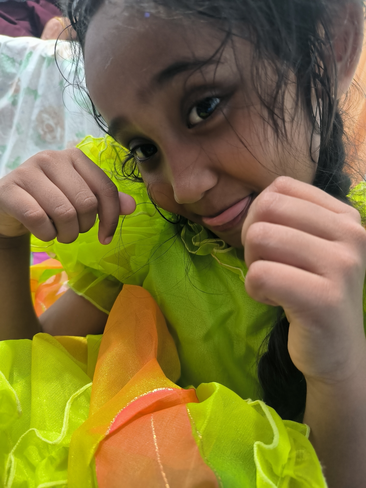 A close-up, eye-level shot captures a young child, likely a girl, from Lhokseumawe, Indonesia, in a playful moment. She looks directly at the camera with one eye while subtly sticking out her tongue, conveying a mischievous or cheeky expression. Her dark, seemingly damp or messy hair frames her face, with individual strands visible.
The child wears a brightly colored, lime-green garment with ruffled details, made of a lightweight, possibly sheer, fabric. Portions of orange and yellow fabric are also visible towards the bottom of the frame, hinting at a vibrant, multi-colored outfit. Her hands are raised and loosely fisted near her face, contributing to the playful posture.
The background is softly blurred, showing hints of a white fabric with a faint pattern and some darker, indistinct shapes, suggesting an indoor or shaded outdoor setting. The lighting appears bright and natural, indicating it is daytime. The overall mood of the image is cheerful and lighthearted. No visible text is present in the image.