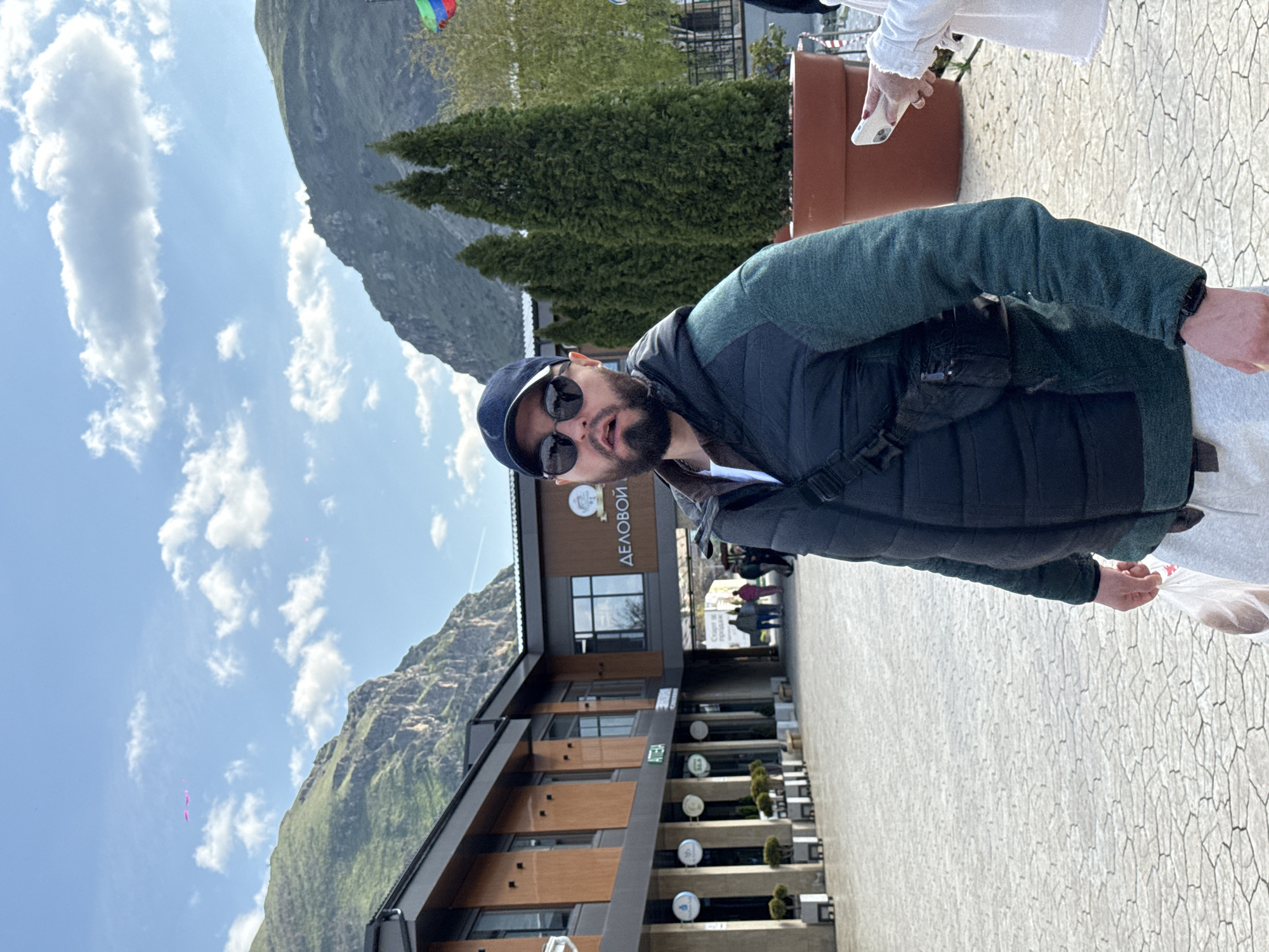 A man stands on a paved walkway in Miatli, Russia. He wears a black cap, sunglasses, a green jacket, and a black vest. Behind him is a two-story building with the word "ДЕЛОВОЙ" visible. Tables with greenery are set up along the building's facade. In the background, there are mountains and a cloudy sky. Other people are walking in the distance near the building. Part of another person wearing a white shirt is visible on the right side of the image.