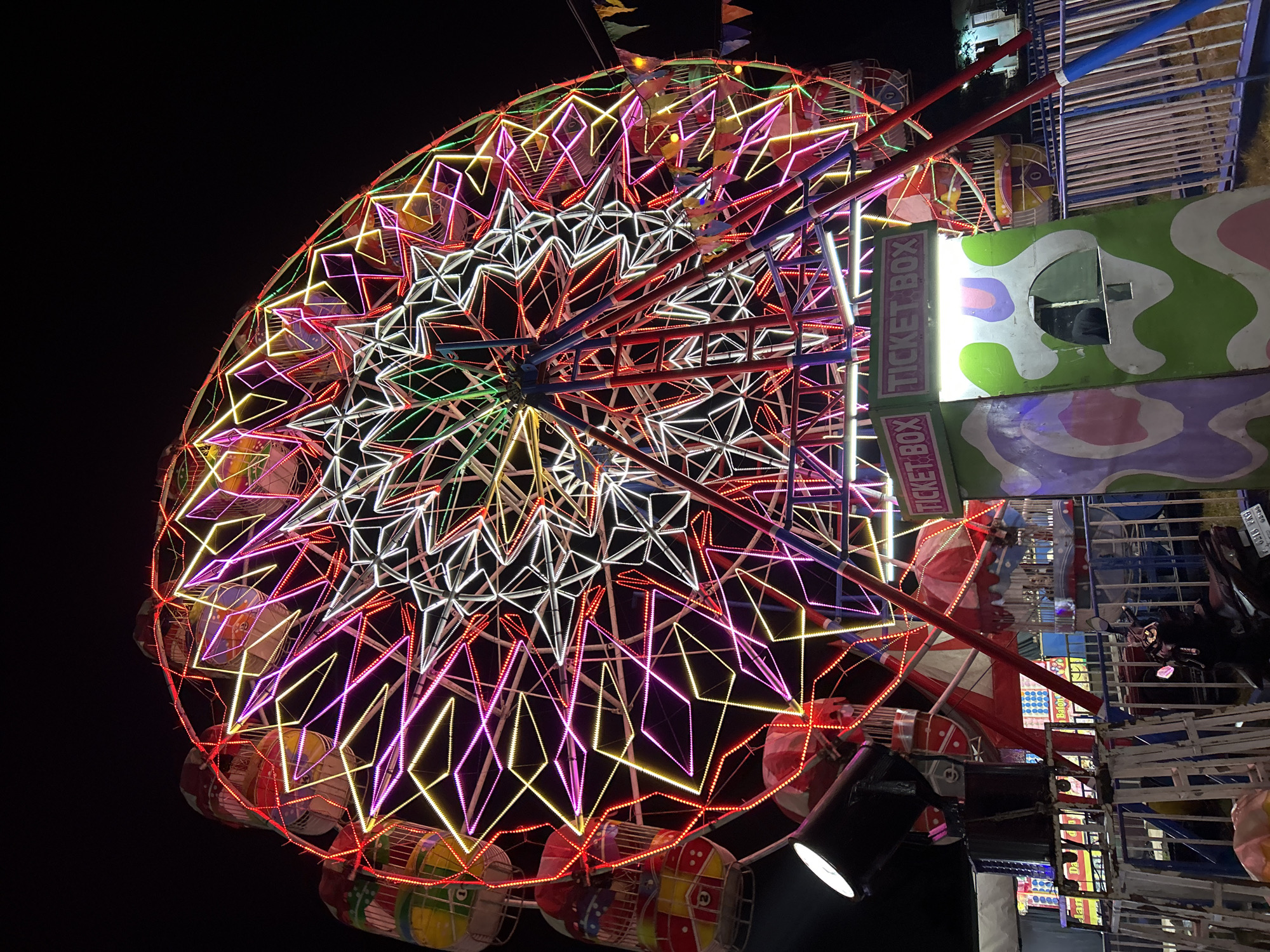 This media file captures a brightly lit Ferris wheel at night, likely at a carnival or amusement park. The Ferris wheel is the primary subject, adorned with an intricate display of colorful lights in white, yellow, pink, and red, creating geometric patterns. Several gondolas are visible, some with vibrant, multi-colored designs.
In the foreground, part of a ticket booth structure is visible, with the text "TICKET BOX" prominently displayed in red lettering on a green and white background. Behind the Ferris wheel, more carnival attractions and structures can be seen, including fencing and other rides, also illuminated with lights. The overall setting appears to be an outdoor event after dark, given the black sky. The image conveys a festive and lively atmosphere typical of a night-time fair. There are no people clearly visible in the image.