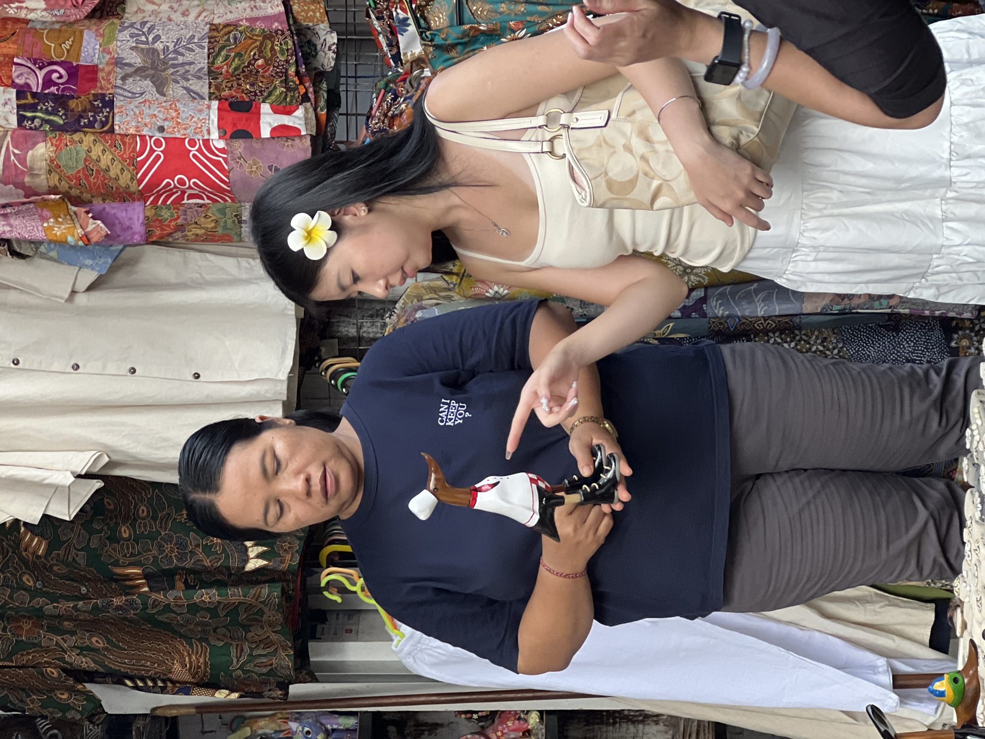 This photograph depicts a vibrant market scene, likely within a stall or small shop in the Ubud District of Indonesia. The image shows two women engaged in an interaction over a decorative item.
On the left, a woman, appearing to be a vendor, holds up a stylized figurine, possibly a traditional Balinese doll or carving. The figurine features a human-like form with brown-toned legs and arms, a white top, and black boots. The vendor wears a navy blue t-shirt with the text "CANI KEEP YOU" printed on it in two lines, and dark gray pants. She looks towards the item with a neutral, perhaps explanatory, expression. A red string bracelet is visible on her right wrist.
On the right, a customer, with a white frangipani flower tucked behind her left ear, points her index finger towards the figurine held by the vendor. She wears a cream-colored spaghetti strap top, a white pleated skirt, and carries a light-colored, patterned handbag over her shoulder. Her left arm is adorned with a smart watch, a silver bracelet, and a light lavender-colored bangle. Her gaze is directed at the item, conveying interest and engagement.
The background is a rich tapestry of folded and hanging textiles, prominently featuring colorful and intricately patterned batik fabrics, indicative of local artistry and craft. The overall setting suggests a bustling, daytime market environment where local goods are sold. The frangipani flower and the style of the textiles and figurine serve as strong cultural cues for the Indonesian location.