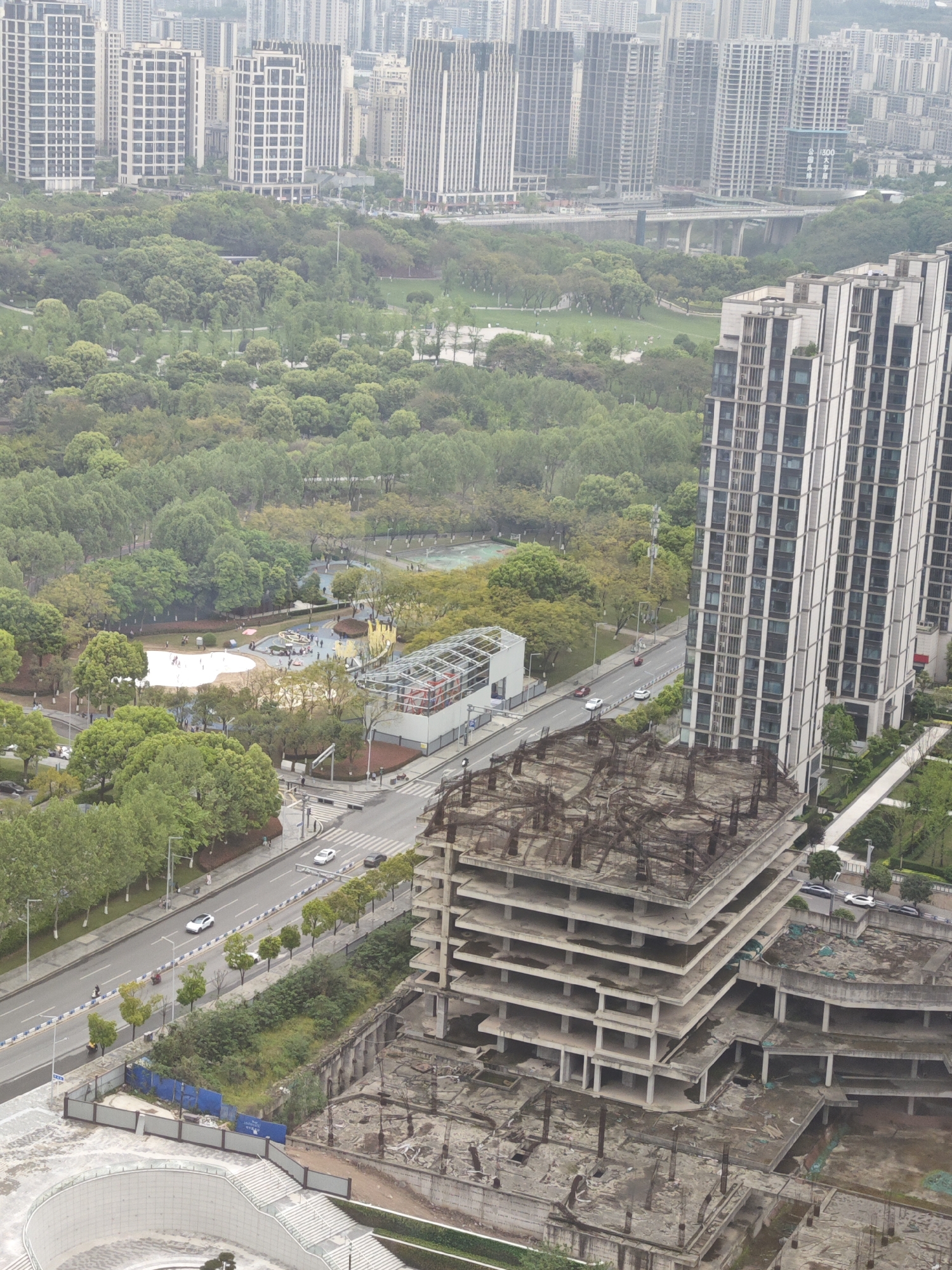 The image depicts an urban scene in Liangjiang, China, characterized by a mix of completed and unfinished construction amidst greenery. In the foreground, a derelict, multi-story concrete building stands in a state of disrepair, with exposed rebar and debris. Adjacent to this structure is a modern, completed high-rise apartment building. A multi-lane road separates these buildings from a park with walking paths, trees, and recreational areas. Numerous people are visible within the park, engaged in various activities. In the background, a dense cluster of high-rise buildings forms the city skyline, partially obscured by a haze. A bridge spans the gap between the built environment and the park area.
