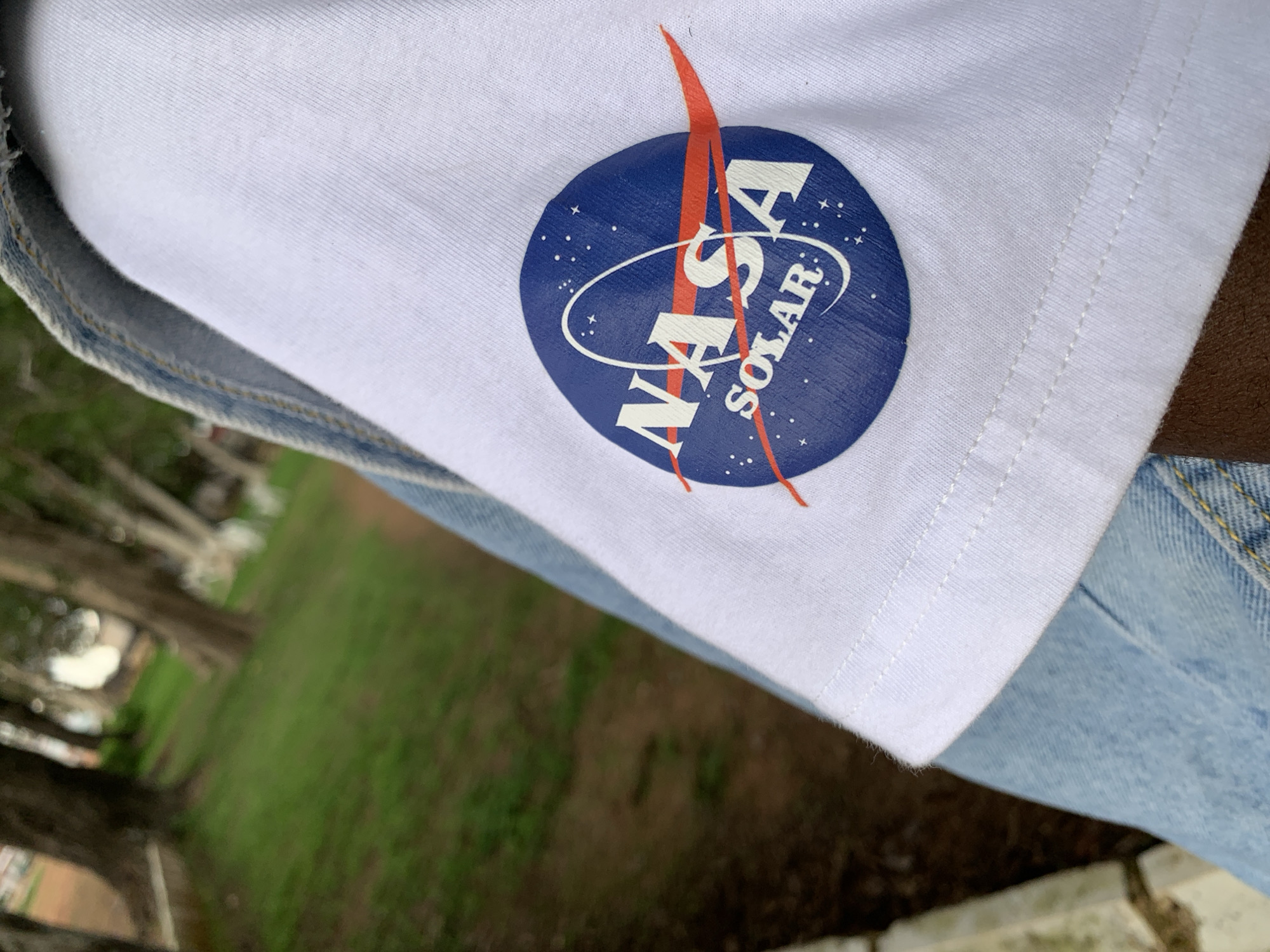 The image shows a close-up of a white t-shirt with the NASA Solar logo. The logo is circular, featuring a blue background with white stars and the word "NASA" in white letters. A red diagonal line cuts across the logo, and below "NASA," the word "SOLAR" is written in smaller white letters. The t-shirt is partially visible, with a portion of light blue denim jeans below it. The background is blurred, showing a grassy area with trees, suggesting an outdoor setting. The lighting appears to be natural daylight, and the overall mood is casual. The image is oriented vertically.