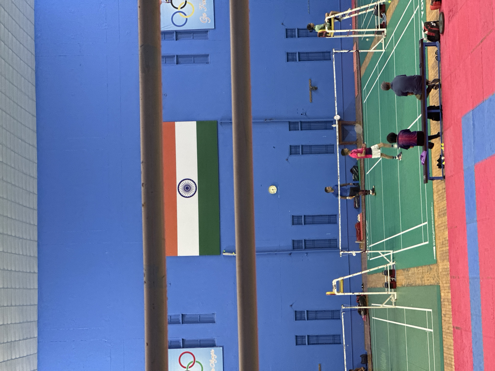 This is a full shot of an indoor badminton court with blue walls. A large Indian flag hangs on the back wall. In the foreground, a metal railing is visible. On the green court, several people are present. A man in a blue shirt stands near a net, and another person in a pink shirt is playing badminton. Two individuals are sitting on a bench in the foreground, and a person is seated on a high chair by the net. Badminton nets are set up on the court, and a clock is visible on the wall. The floor is a mix of green court surfaces and red and blue mats.
