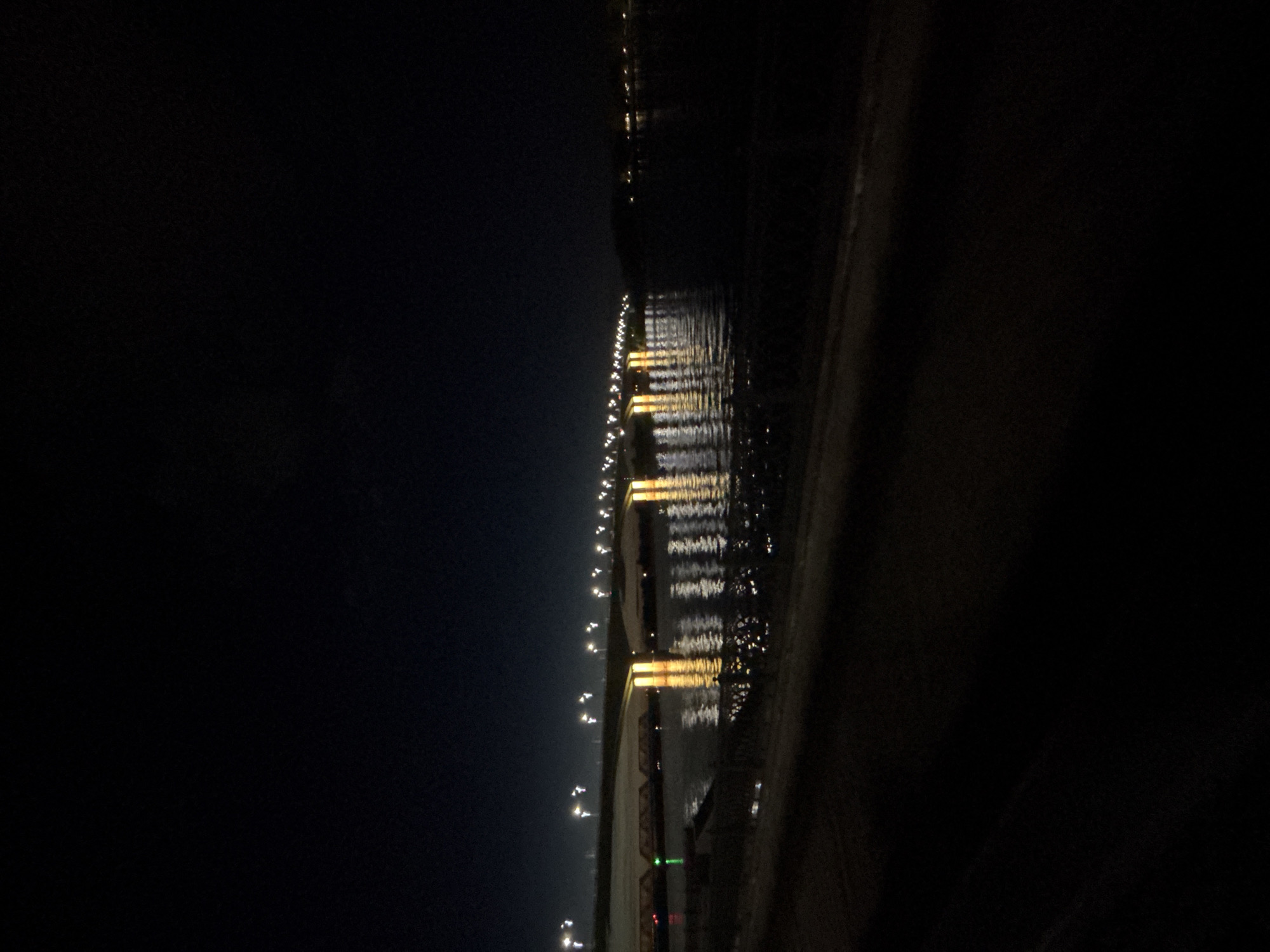 The image depicts a night scene in Rusanovo, Russia, showing a row of streetlights along a body of water. The lights are reflected on the water's surface, creating a shimmering effect. The sky is dark, suggesting it is nighttime. In the distance, more lights are visible on the opposite shore. The immediate foreground appears to be a darker area, possibly a shoreline or embankment.