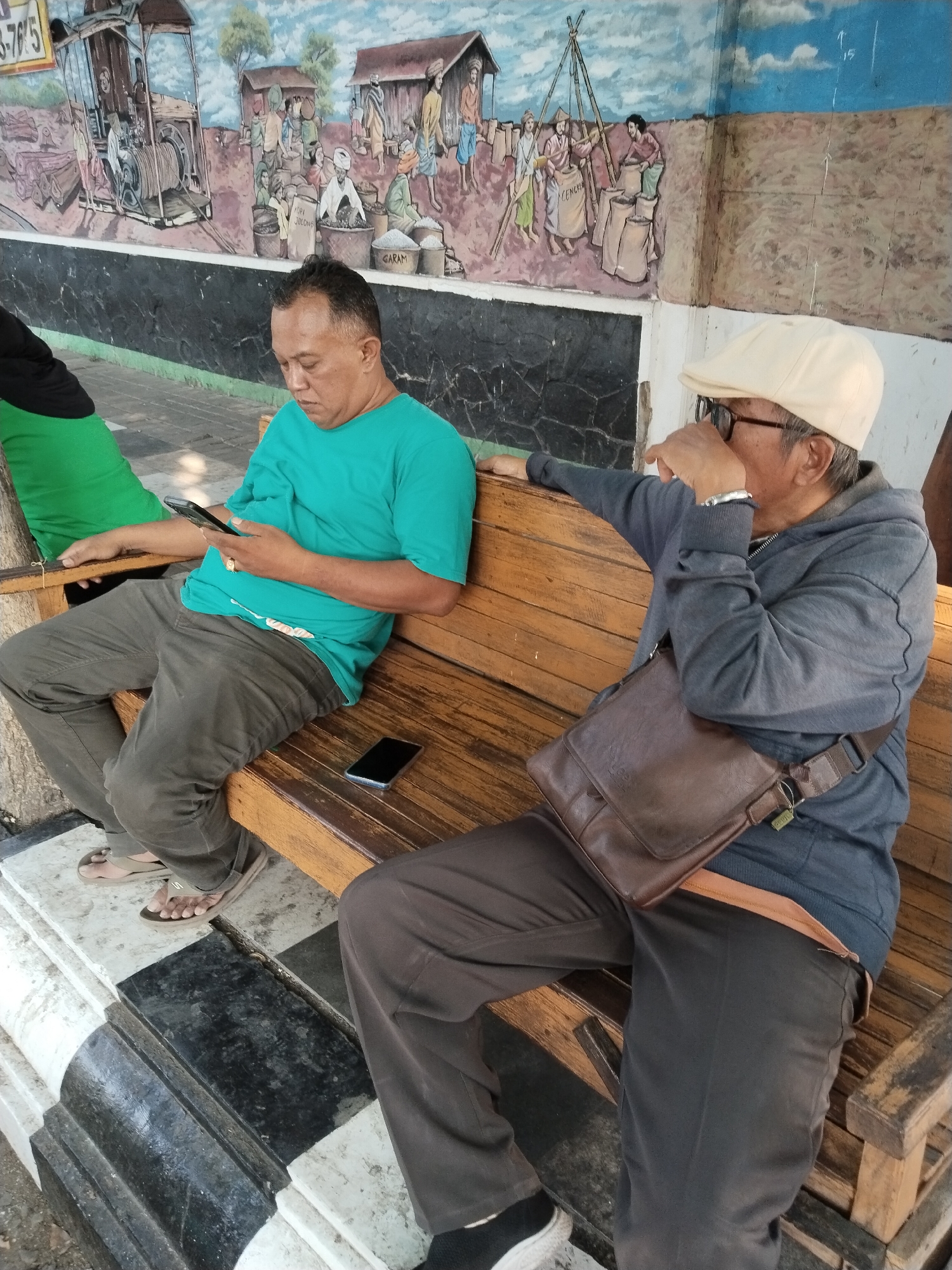 Two men are seated on a wooden bench in front of a mural depicting a village scene with a train and agricultural activities. The man on the left, wearing a turquoise t-shirt and grey pants, is looking at his smartphone. He has short, dark hair and is wearing flip-flops. The man on the right, wearing a grey hoodie and dark pants, has a beige cap on and glasses. He is holding his hand to his face, appearing to be thinking or resting. A brown leather messenger bag is slung across his body. A black smartphone is placed on the bench between the two men. The bench is positioned on a patterned tiled area, with a black and white pattern at the base. The mural behind them is painted on a wall and depicts a historical or traditional Indonesian setting. The overall environment suggests an outdoor waiting area, possibly in a public space within Kecamatan Pati, Indonesia, given the mural's context.