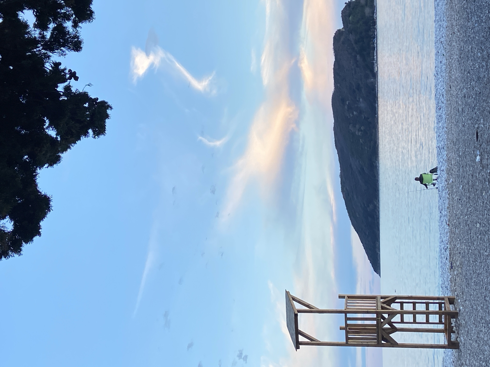 The scene is a coastal view in or near Budva, Montenegro. The foreground shows a sandy or pebbly beach with calm, reflective water. A wooden structure, possibly a lifeguard stand, is situated on the beach. In the distance, a silhouette of a mountain rises from the water, suggesting a bay or inlet. A person is visible on the beach near the water's edge, though details are limited. The sky is predominantly blue, with streaks of white clouds tinged with orange or yellow, indicating either sunrise or sunset. A tree's branches are visible in the upper left corner of the frame.