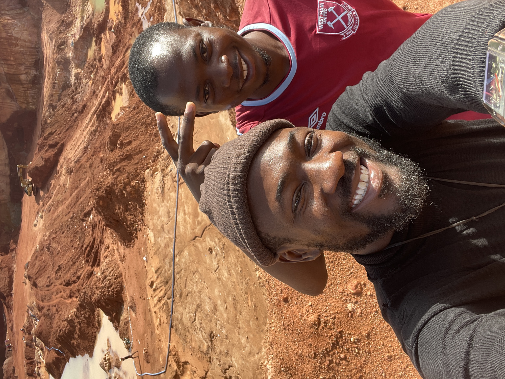 Two men are posing for a selfie in Kwang, Nigeria. The foreground shows the man taking the selfie wearing a brown beanie and a black turtleneck sweater, displaying a peace sign. Behind him is another man wearing a West Ham United FC shirt. The background reveals a large excavated pit in the ground, indicating a mining site. A small excavator is visible in the distance. The ground around the pit is primarily reddish-brown soil, and there's some water pooled in the lower areas of the excavation.