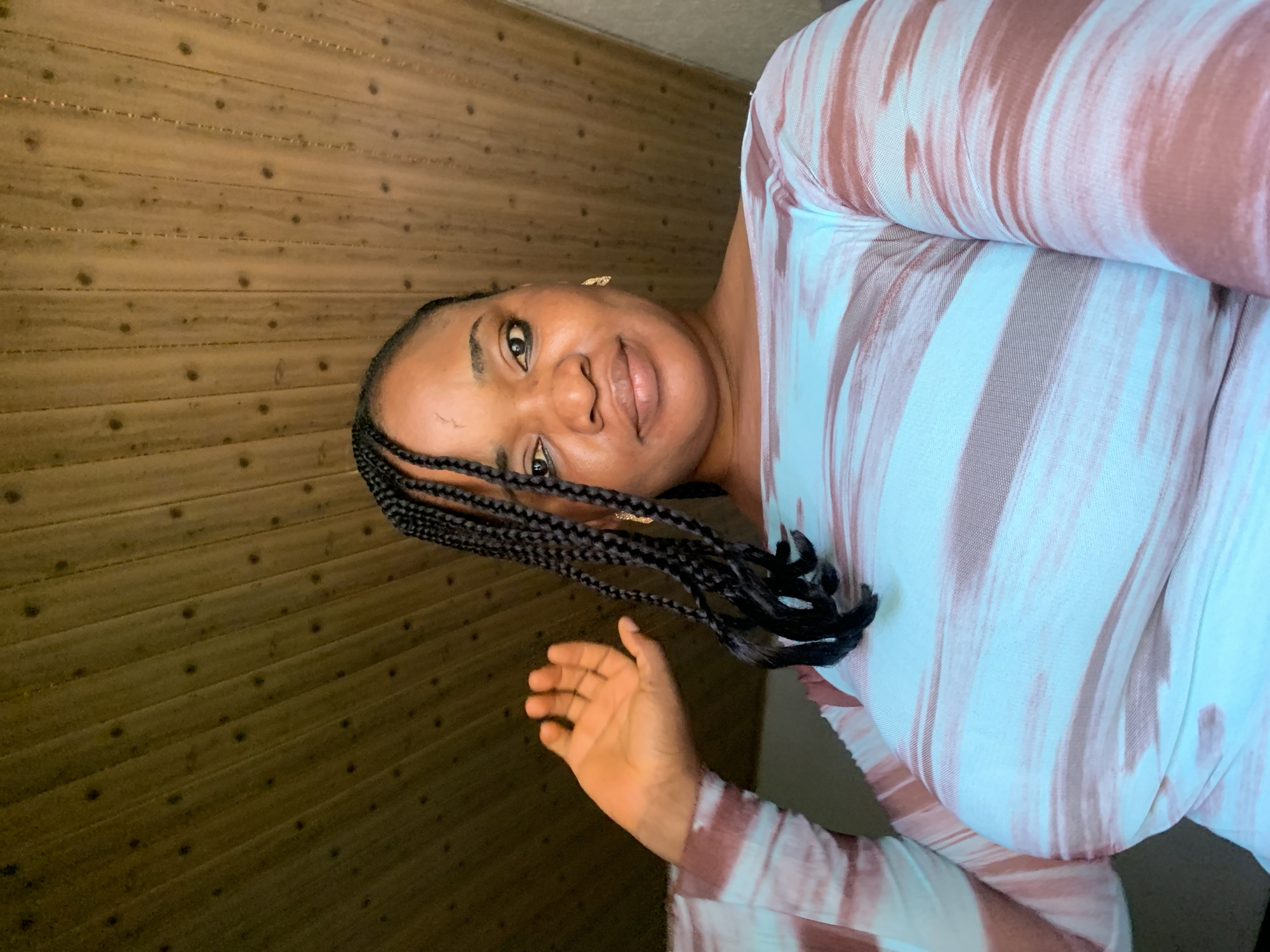 A woman with dark braided hair and light brown skin is looking at the camera. She is wearing a light blue and brown striped long-sleeved shirt. Her left hand is raised and open, with her palm facing the camera. She is indoors, in front of a wooden wall. The setting is likely a residential interior.