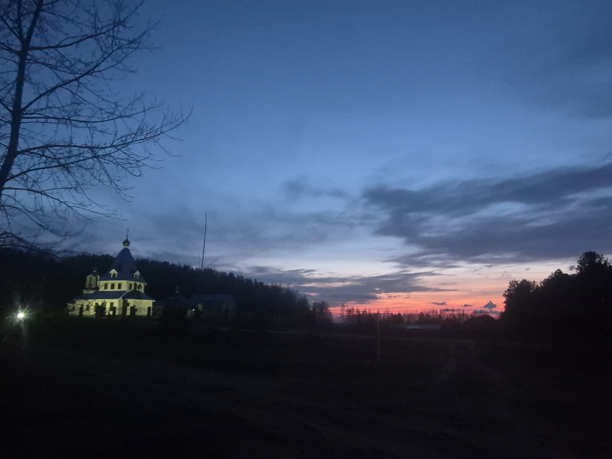 This media file captures a serene twilight landscape in Chaikovsky, Russia, at dusk or dawn. The scene is dominated by a dramatic sky and a distinctive church building.
In the foreground, a prominent bare tree with dark, intricate branches occupies the left side, silhouetted against the sky. The immediate ground is dark and unlit, likely a field or unpaved area. A bright artificial light source, possibly a streetlamp, emits a distinct starburst glow near the bottom left corner.
The mid-ground features a white or light-colored church structure, illuminated by warm artificial light, standing out against a dark forested hill behind it. The church exhibits typical Eastern Orthodox architecture with a central dome, smaller decorative spires, and multiple windowed levels.
The sky is a key element, displaying a striking gradient of colors. The upper portion is a deep, dark blue, transitioning downwards through lighter blues and grays with scattered clouds. Towards the horizon on the right, a vibrant band of orange and red indicates either a sunrise or sunset, contrasting sharply with the cooler tones above. Dark silhouettes of distant trees line the horizon on the right.
The overall atmosphere is tranquil and contemplative, with no discernible human activity. The artificial lighting on the church creates a focal point against the darkening natural environment. No text is visible in the image.