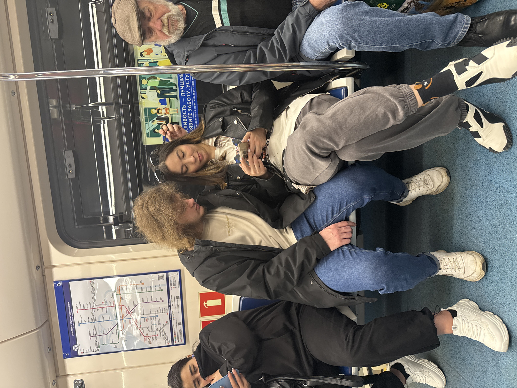 Here is a description of the image:
**Scene:** The image captures the interior of a subway car, likely in Saint Petersburg, Russia, given the map visible on the wall.
**People:**
* There are at least four individuals visible.
* An older man with a gray beard and a hat is sitting.
* A woman in a black leather jacket is looking at her smartphone.
* A man with long curly hair is sitting next to her.
* Another person is looking at a smartphone.
**Objects:**
* Subway map
* Mobile phones
**Actions:**
* People are sitting in the subway car.
* Some individuals are using their phones.
**Environment:**
* The interior of a subway car, evident from the seats, walls, and subway map.