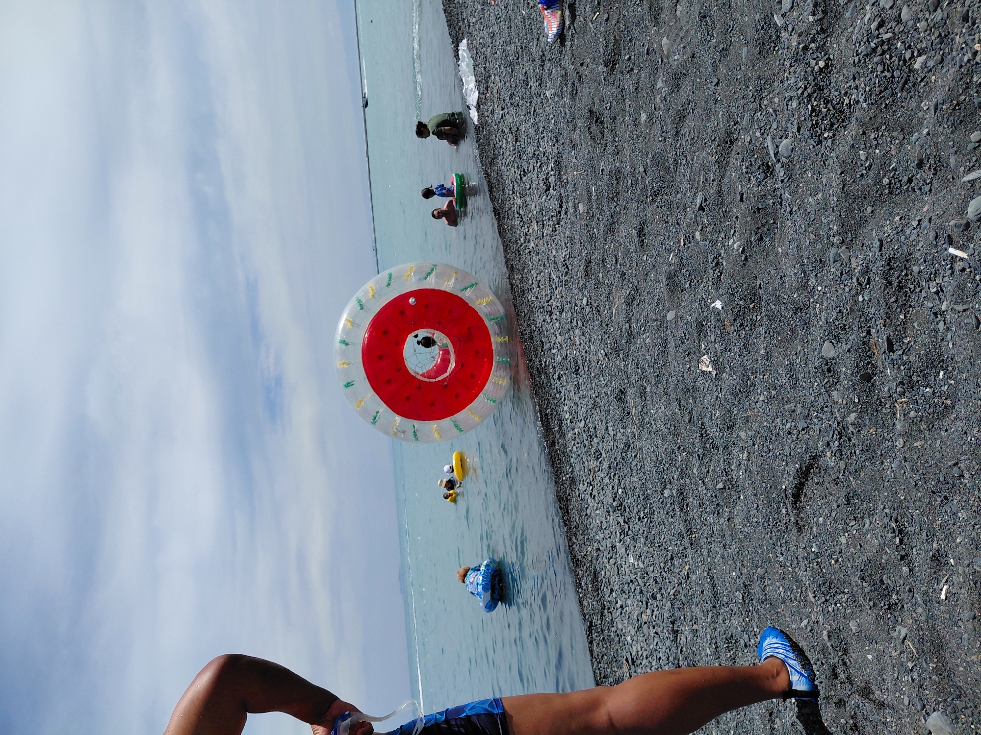 In this image, taken at a lakeside beach in the vicinity of Ақши, Kazakhstan, a group of people are engaged in water activities. A large, transparent inflatable sphere with red and yellow accents is prominently positioned in the water, with a person visible inside. Several individuals are scattered in the shallow water, some in inflatable rings. The foreground features a person's legs and feet clad in blue and white water shoes, standing on a dark, pebble-strewn beach. The water is a light blue-green, and the sky above is partly cloudy with a distant boat on the horizon.
