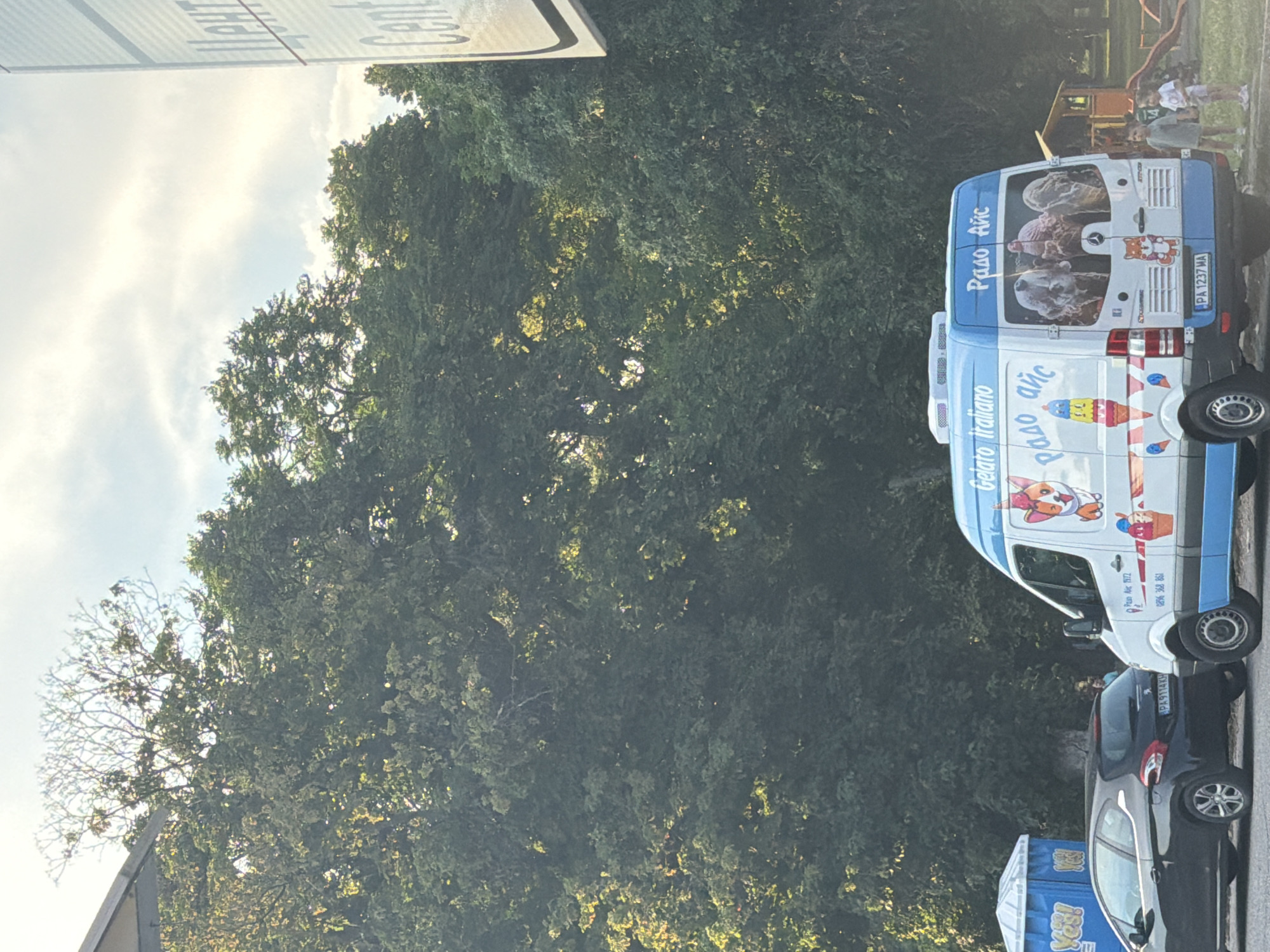 The image shows a street scene in the city of Pazardzhik, Bulgaria. In the foreground, an ice cream van, branded "Gelato italiano" and "Радо айс", is parked. The van is decorated with cartoon images of ice cream and a fox. Beside the van, two children, a boy and a girl, are standing, possibly waiting for ice cream. To the left of the ice cream van, a black car is parked next to two portable toilets, one blue and one white, with "Yes!" written on them. In the background, lush green trees dominate the scene, with a partly cloudy sky overhead. A sign with lettering in what appears to be Cyrillic and Latin alphabets is visible on the far right, with the word "Center" visible in Latin. A children's playground with a slide can be seen behind the ice cream van. The overall setting suggests a public area within the city, likely a park or a recreational zone, during daytime.