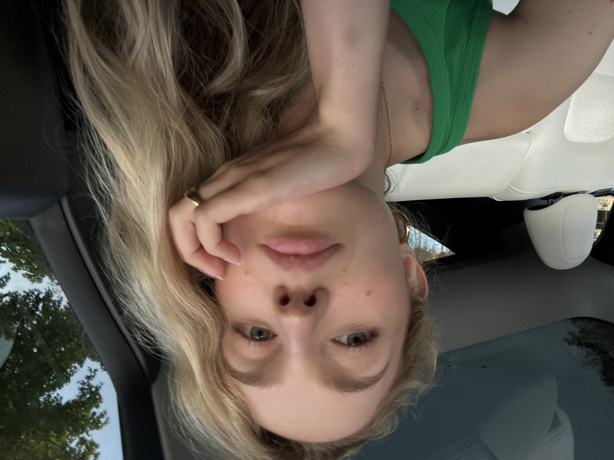 A young woman with blonde hair is sitting in the passenger seat of a car. She is wearing a green tank top and gold jewelry, including a hoop earring and a ring. Her hand is resting on her chin, and she is looking at the camera. The car has white leather seats and a sunroof, suggesting it is a luxury vehicle. The background shows trees and a clear sky, indicating an outdoor setting, possibly in a suburban or park area within Coral Gables, Florida, United States of America. The image appears to be a selfie.