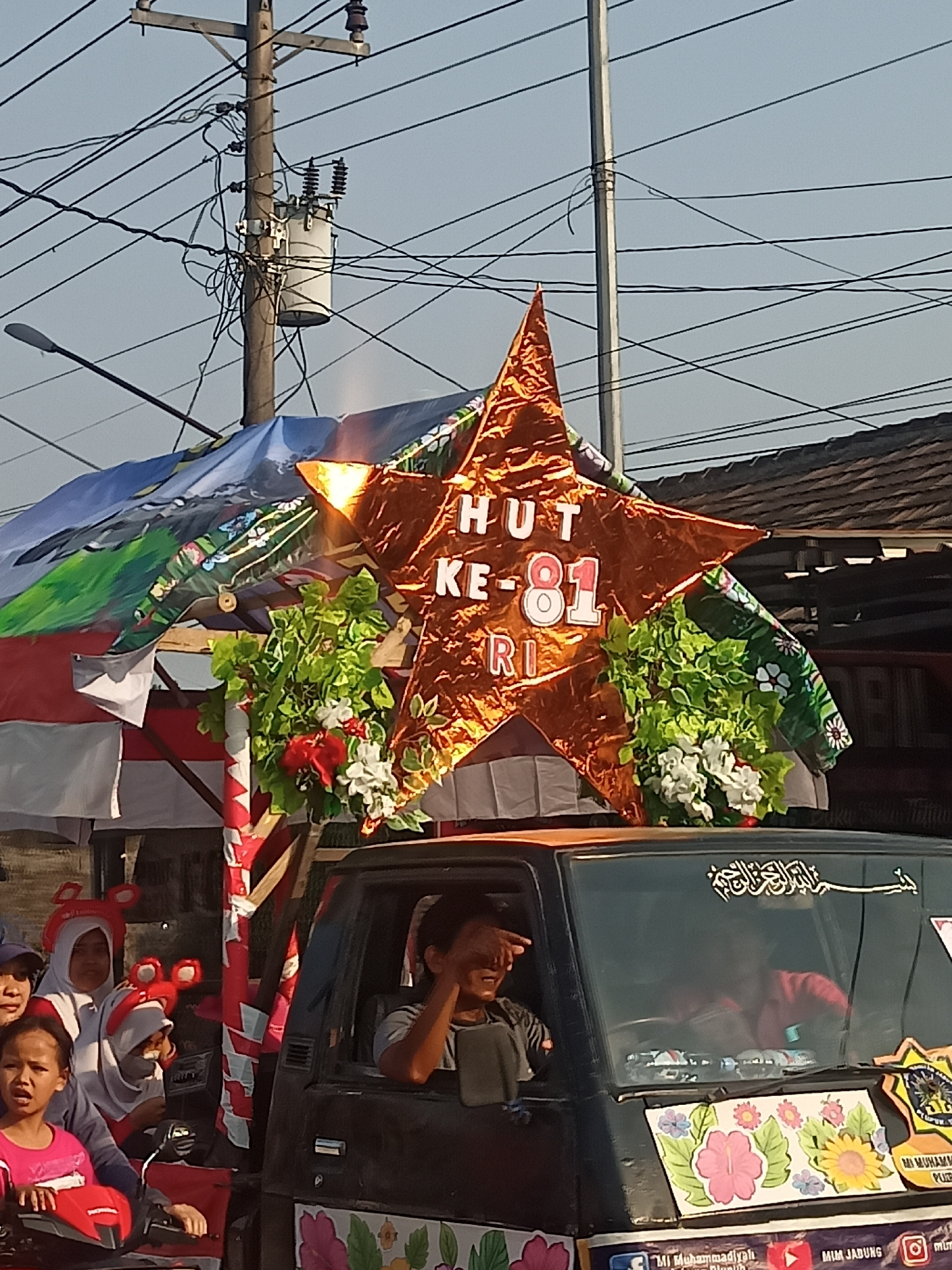 The image depicts a parade float in Ngrompongan, Indonesia, celebrating "HUT KE-81 RI" which signifies the 81st anniversary of the Republic of Indonesia. The float is adorned with a large, orange, star-shaped decoration featuring the text "HUT KE-81 RI" in white lettering. Green foliage and white flowers are arranged around the star, adding to the festive appearance. The float appears to be mounted on a dark-colored vehicle, likely a van or truck, with several people visible. A man is driving the vehicle, looking to his right. In the back of the vehicle, several children are seen, some wearing headwear with animal ear designs. The scene is set against a backdrop of utility poles, electrical wires, and buildings, suggesting an urban or semi-urban environment under a clear sky.