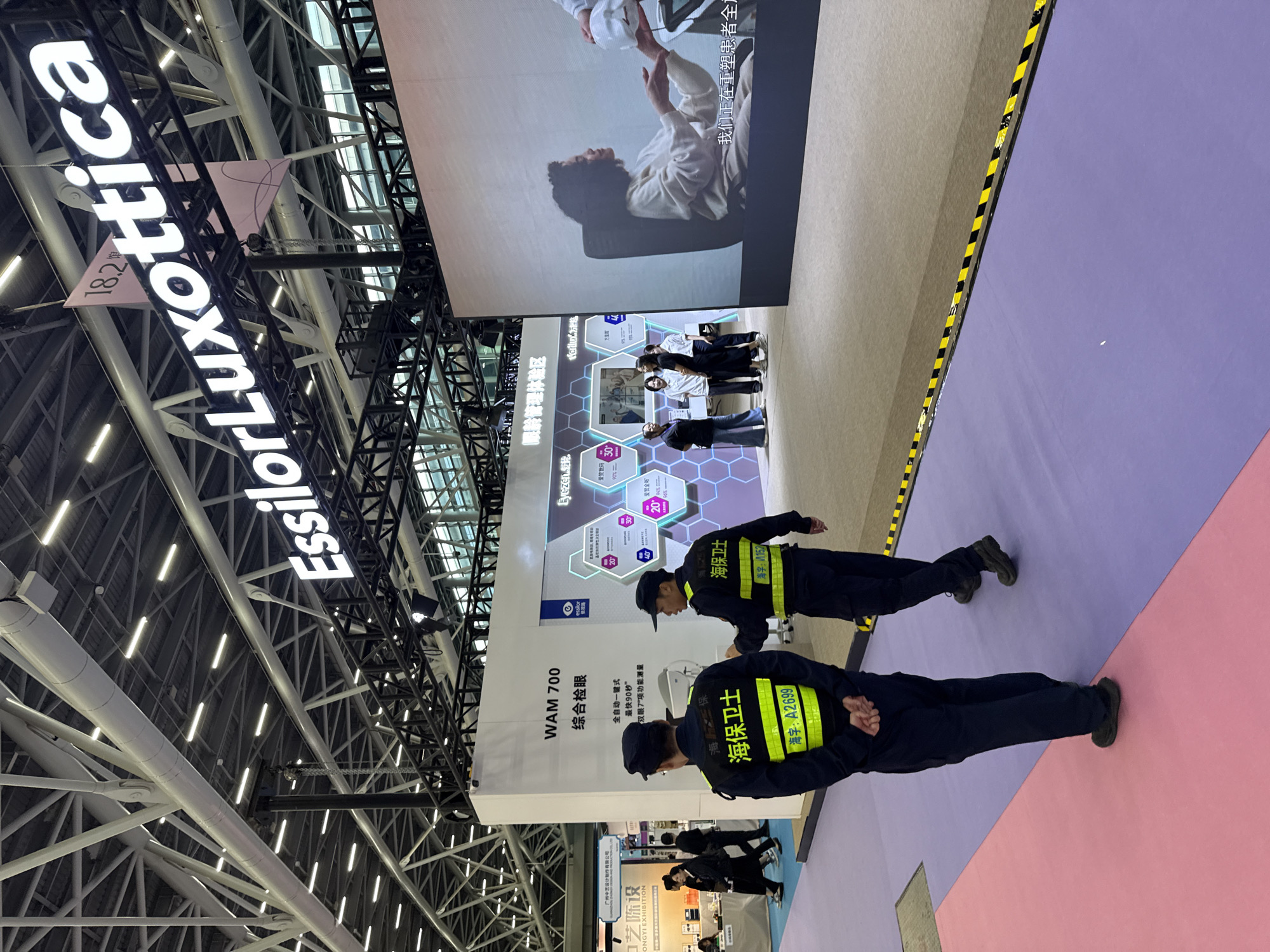 The image depicts an indoor exhibition hall, likely at the Guangzhou International Lighting Exhibition held in Haizhu District, China, based on the presence of event signage and the general ambiance of a trade show. Dominating the upper portion of the image is a large, illuminated sign reading "EssilorLuxottica," a prominent eyewear company. In the foreground, two individuals in dark blue security uniforms with bright yellow reflective vests stand facing away from the camera. Their vests bear the text "海保卫士" (Hǎi bǎo wèishì, meaning "Sea Guard" or "Maritime Guard") and numerical designations. The individual on the left has his hands clasped behind his back, while the one on the right appears to be gesturing or speaking to the other.
Behind the security personnel, a large white booth displays information about "WAM 700 综合眼检" (WAM 700 Comprehensive Eye Examination). The booth features graphics and text related to eye care technology. To the right of this booth is a massive screen displaying a video or image of a woman interacting with what appears to be eye examination equipment, with Chinese text overlaid on the bottom of the screen. Several other individuals are visible in the background, interacting at various booths or walking through the exhibition space. The floor is a patchwork of colors, with sections of pink and purple carpet separated by a yellow and black striped caution tape. The overall environment suggests a professional and bustling event.