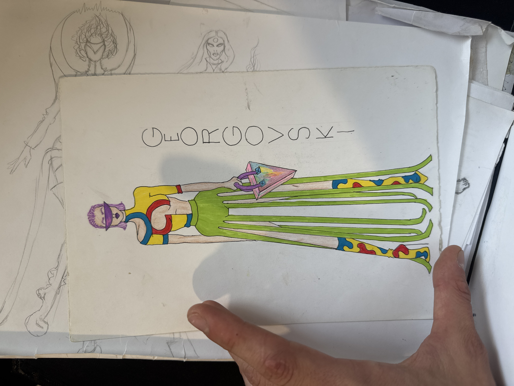 This media file shows a drawing of a stylized figure with purple hair and a visor, wearing a colorful outfit consisting of a yellow top with red and blue accents, a green skirt, and patterned leggings. The figure has long, green, tentacle-like appendages extending from its waist. In its hands, the figure holds a triangular object with a rainbow gradient and colored studs, grasped by purple straps. Above the figure, handwritten text in black ink reads "GEORGOVSKY-". The drawing is on a sheet of white paper, which is layered over other papers and sketches. To the left of the drawing, faint pencil sketches of figures and what appear to be winged creatures can be seen on underlying sheets. A thumb and finger are visible in the foreground, partially obscuring the bottom right of the image. The drawing style is illustrative and cartoonish. The setting is likely an artist's studio or workspace, given the presence of multiple sketches and papers. No specific time of day, weather, or location cues within Sofia, Bulgaria are evident in the artwork itself. The overall emotion conveyed by the artwork is difficult to ascertain due to its stylized nature, but the bright colors might suggest a playful or energetic mood.
