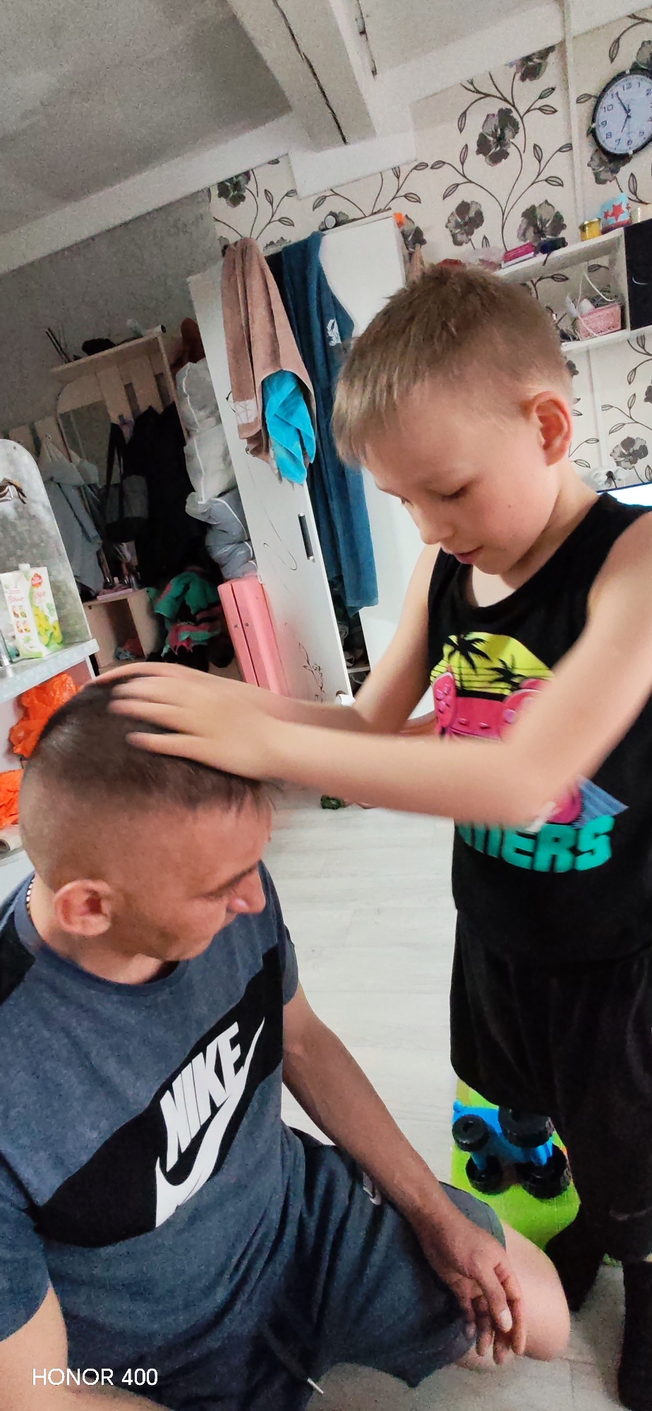 This indoor image from Пушкари, Russia, features an adult male and a young boy. The man, sporting a very short, faded haircut, is seated and wears a grey t-shirt with a prominent black and white "NIKE" logo, along with dark shorts. The boy, with light brown hair, stands beside him, dressed in a black tank top with a colorful graphic design that includes the partial text "MERS." He has both hands gently placed on the man's head, appearing to be giving a massage or playfully interacting with his hair.
The setting is an indoor space, likely a home, with patterned wallpaper featuring dark floral motifs. In the background, a white cabinet or shelving unit is visible, holding various items, and clothes are hanging, including a brown towel and blue fabric. A round wall clock is mounted on the wall to the upper right. On the floor near the boy's feet, a bright green and blue toy car can be seen. The ambient lighting suggests it is daytime. The man appears focused downwards, while the boy seems engaged in his activity. A "HONOR 400" watermark is present in the bottom left corner of the image.