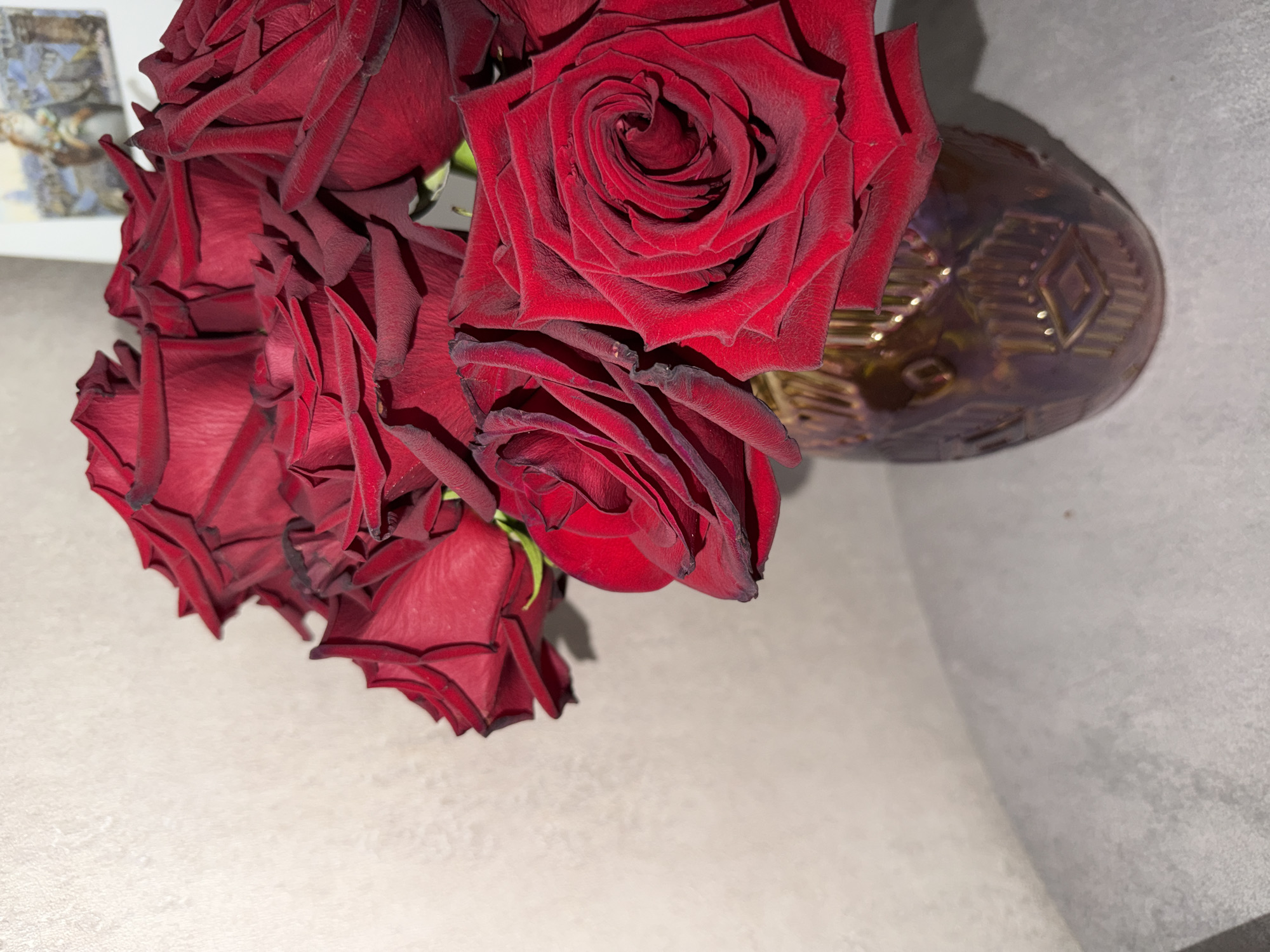 A close-up, low-angle shot showcases a bouquet of deep red roses in a decorative, iridescent brown vase. The roses are densely packed, with some fully bloomed and others still in bud, their velvety petals slightly wilted at the edges. The vase has an intricate embossed pattern of vertical lines and diamond shapes, reflecting light with a metallic sheen. The background is a plain, textured off-white wall, with a faint, out-of-focus image of a framed picture visible on the upper right. The arrangement sits on a textured gray surface. The image is taken indoors, likely in a home setting, with soft lighting casting shadows. There are no people or animals visible. The scene is devoid of any specific geographical markers that would indicate its location in Petropavlovsk-Kamchatsky, Russia, or any other city.