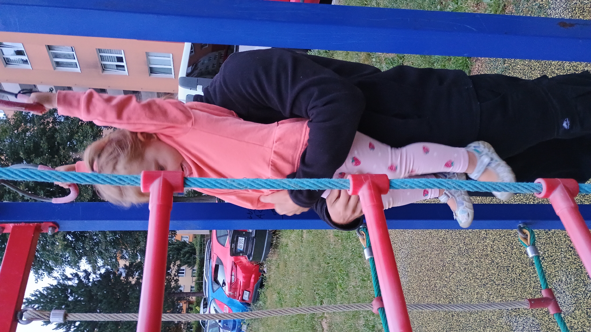 In Sokolov, Czechia, a young girl with blonde hair pulled back is being held aloft by an adult, who is wearing a black hooded sweatshirt and black pants. The girl is wearing a pink long-sleeved shirt and pink leggings adorned with a strawberry pattern. She has on light-colored shoes. They are on a playground with blue and red climbing structures. Ropes are visible, one blue and one grey. In the background, cars are parked, and a multi-story apartment building stands. The ground surface of the playground is a mottled yellow and black material. The overall lighting suggests it is daytime.